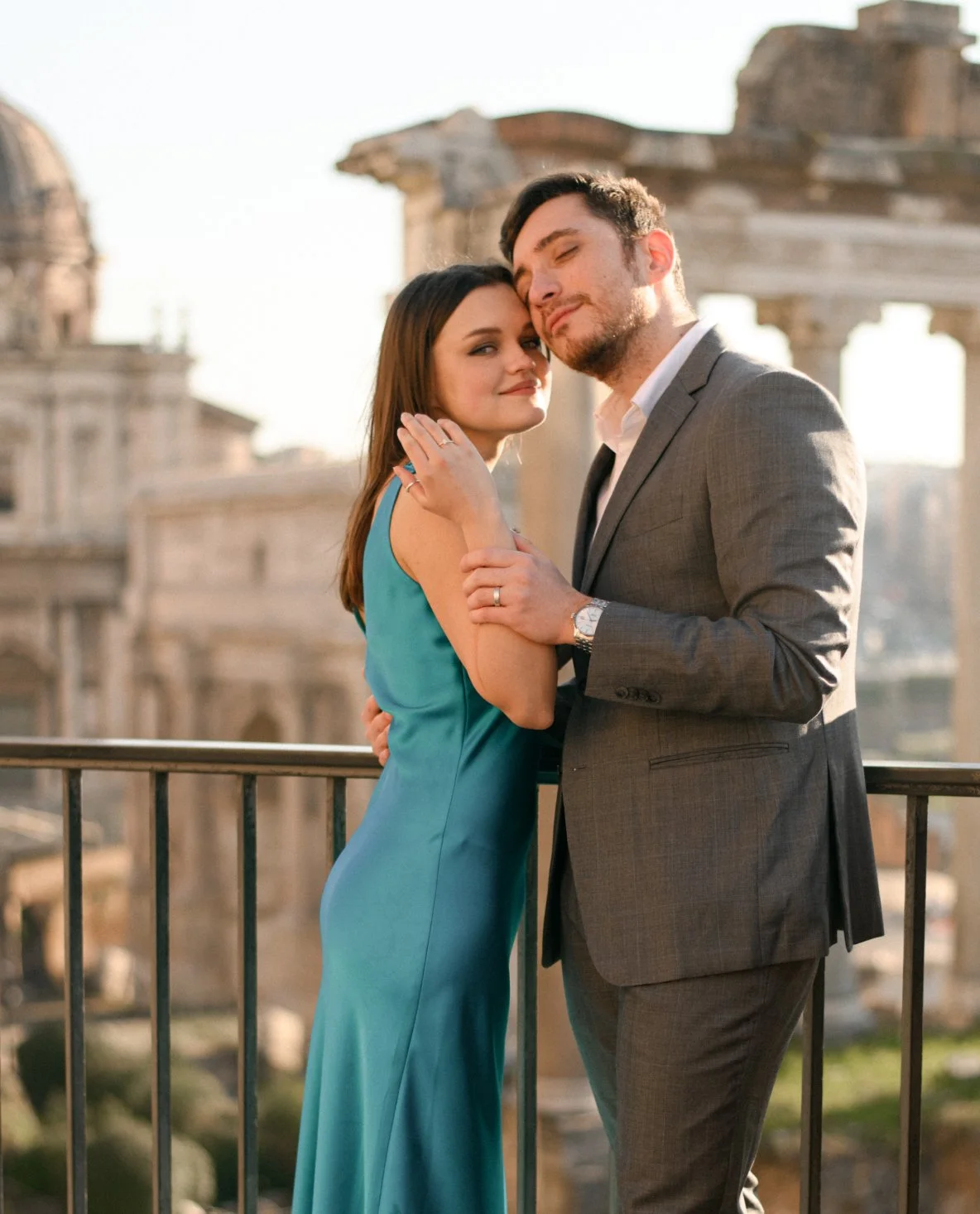 A romantic couple standing close on a balcony with ancient ruins in the background, both in formal attire, sharing an intimate moment.
