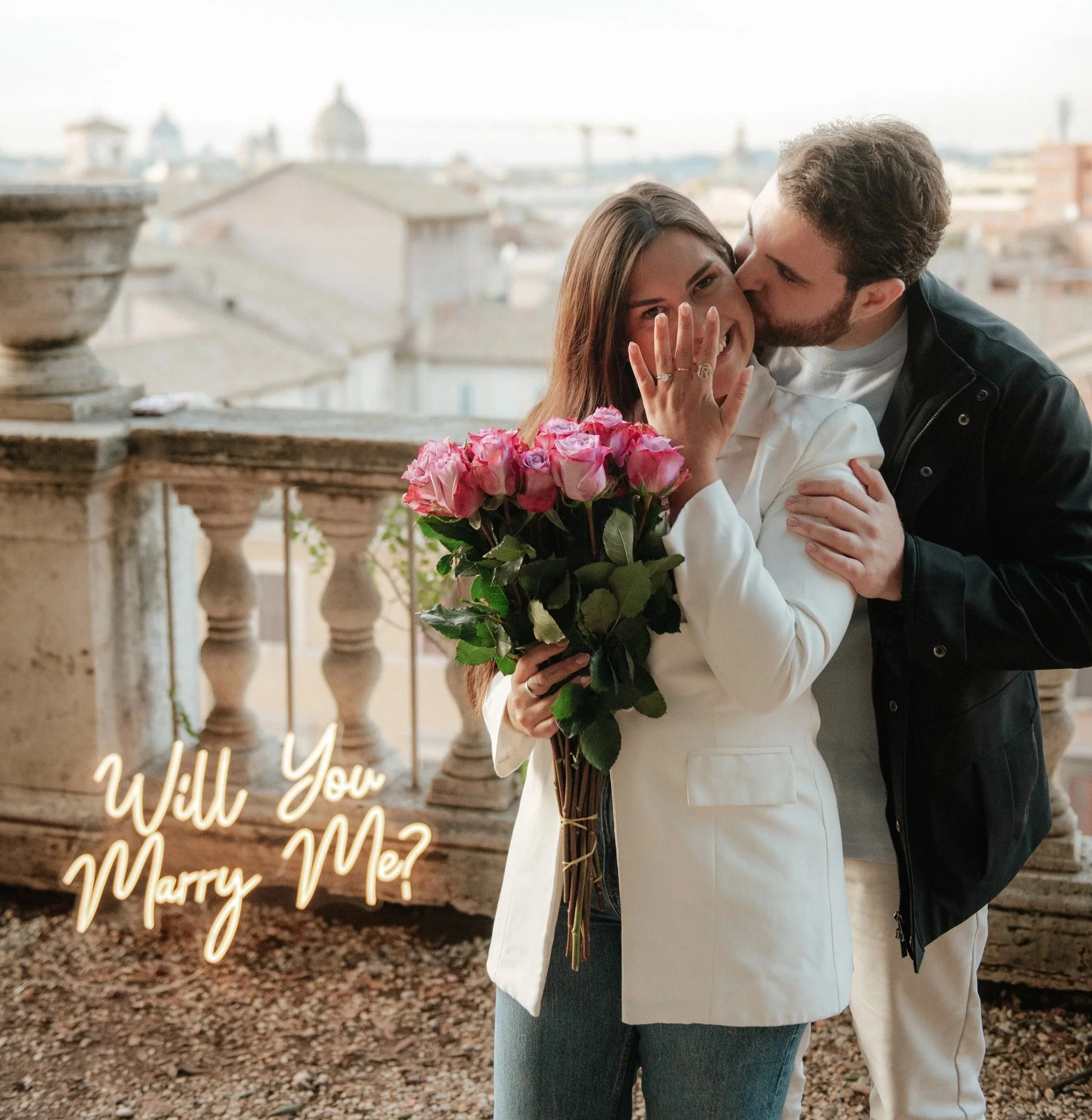 A man kisses a woman on the cheek while she holds a bouquet of pink roses. The woman smiles in a white jacket, and the man wears a black jacket. The background shows a cityscape with rooftops and a cloudy sky. There is a glowing text overlay that says, 'Will You Marry Me?'