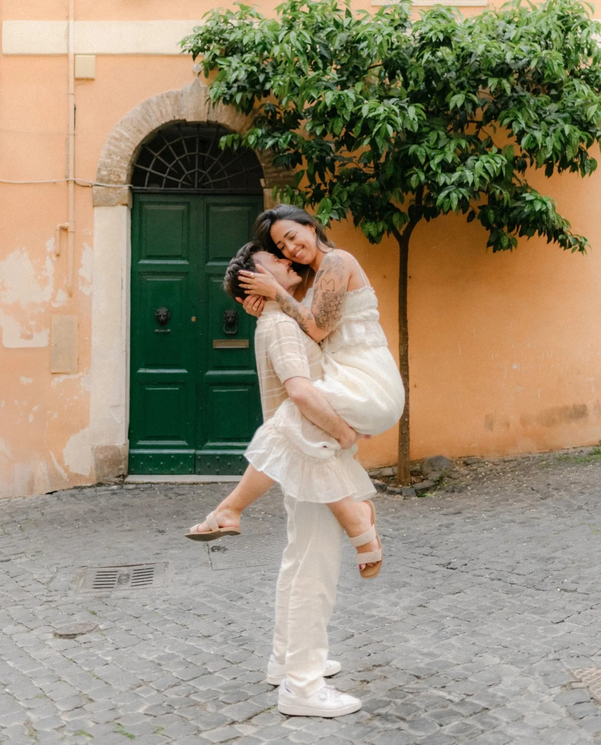 A man lifts a woman in his arms while they smile and embrace on a cobblestone street. A green door and an orange wall behind them, with a tree to the right.