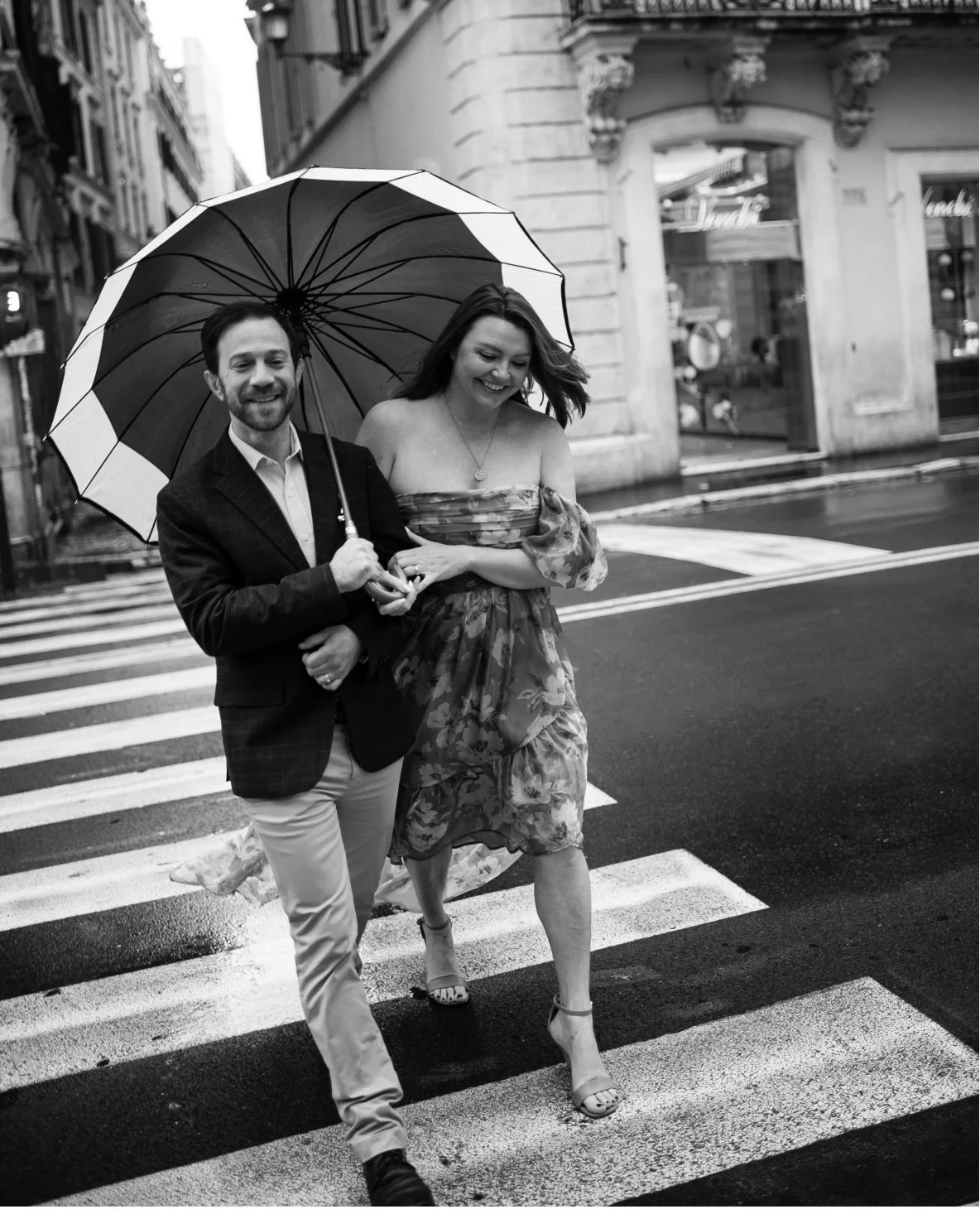 A happy couple walking arm-in-arm on a city crosswalk while holding an umbrella, smiling and enjoying moment together in stormy weather, in a black and white photo.