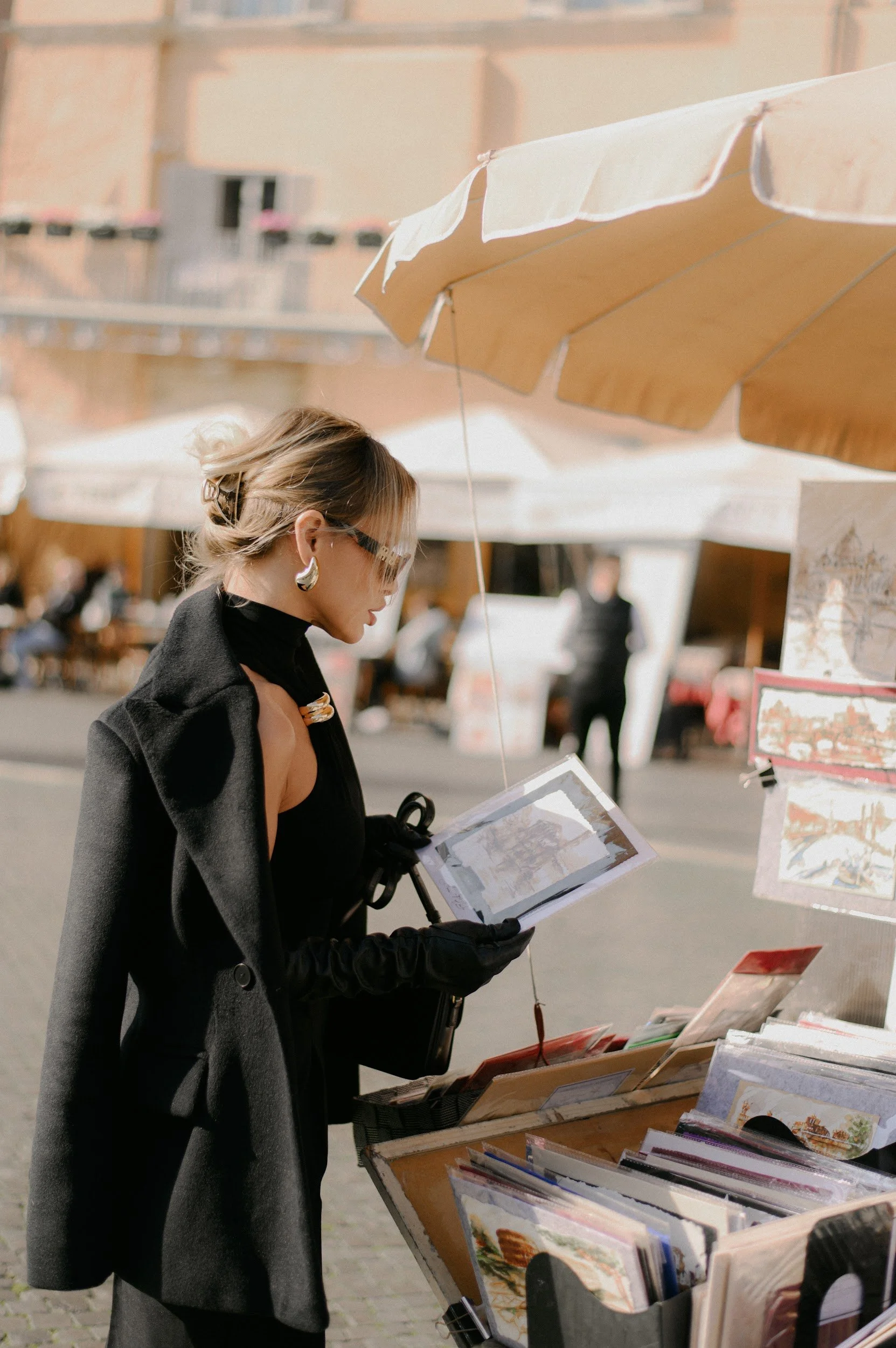 A woman with short blonde hair, wearing glasses, a black turtleneck, a dark coat, black gloves, and earrings, is browsing books and artwork displayed on a stand at an outdoor marketplace at Piazza Navona