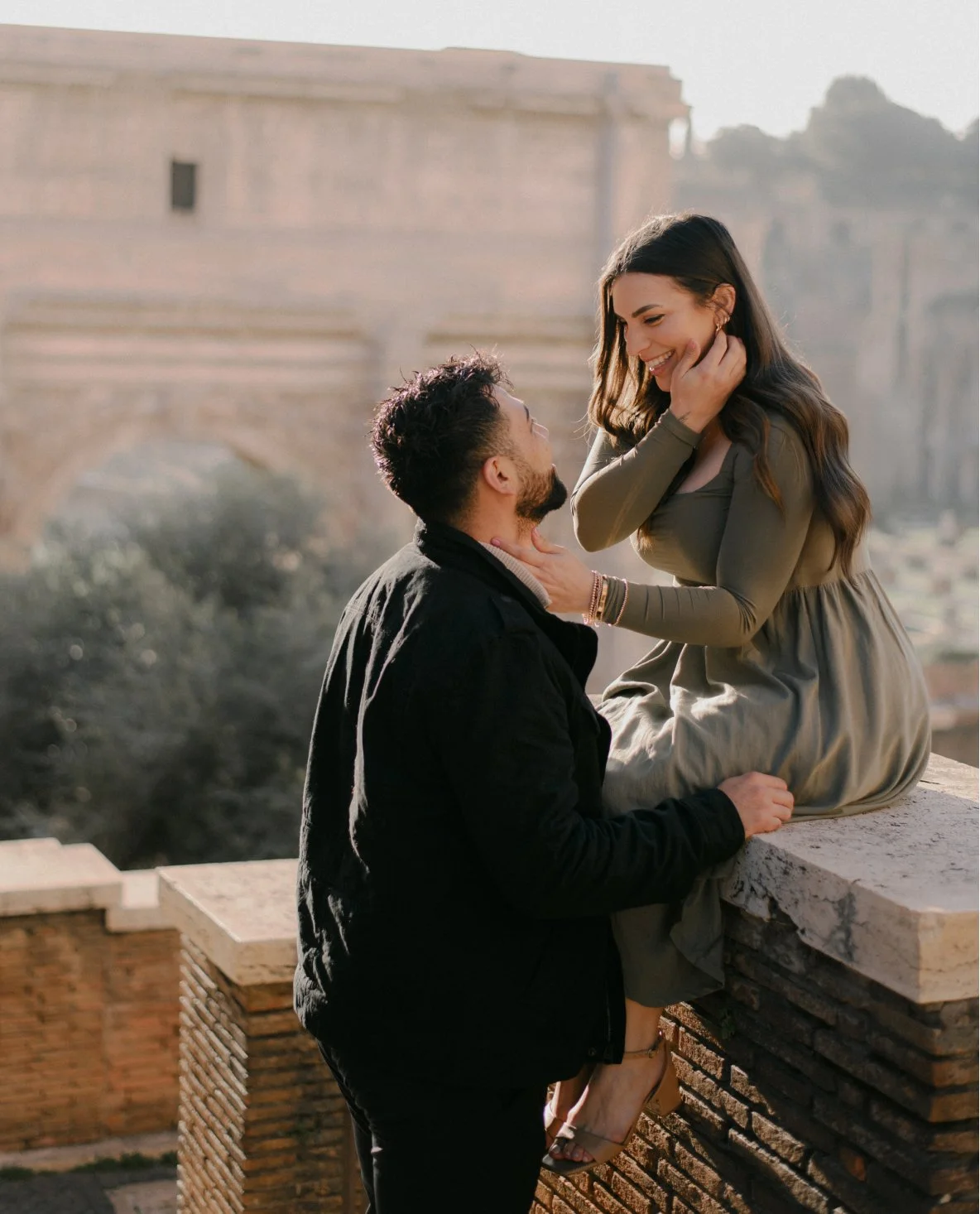 A couple sharing an intimate moment outdoors, with the woman sitting on a ledge and the man kneeling before her, both smiling affectionately.