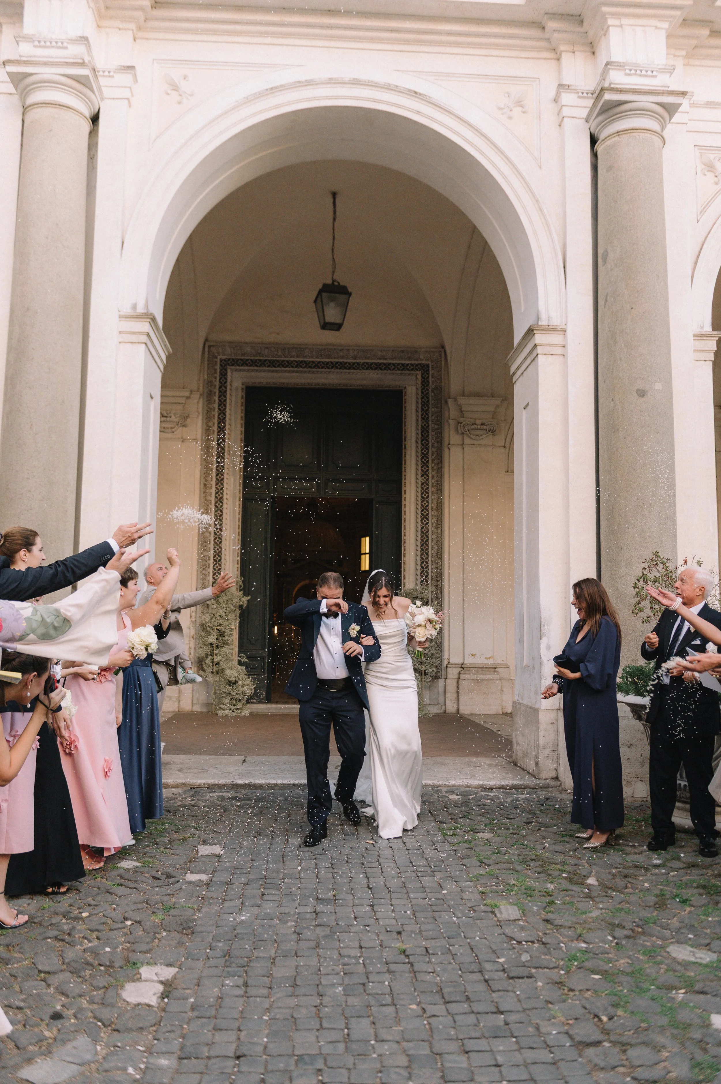 Newlywed couple walking out of a church in Rome, surrounded by friends and family throwing rice or confetti as they celebrate their wedding.