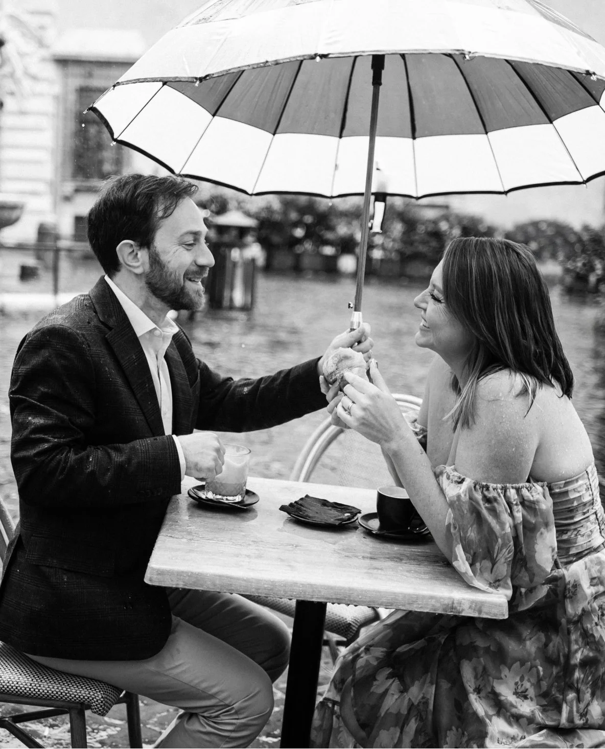 A black and white photo of a man and woman sitting at an outdoor table by a lake, sharing a moment under an umbrella in the rain. The man is smiling and handing the woman a sandwich, while the woman, also smiling, is reaching for it.