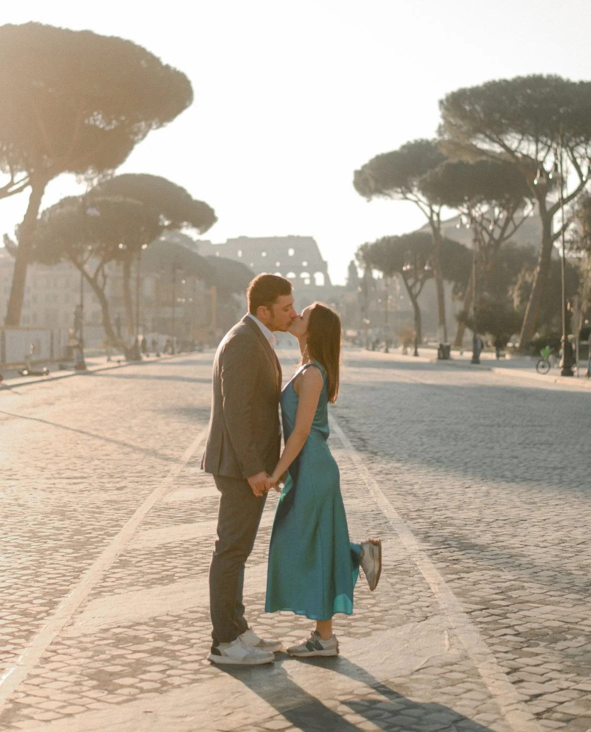 A couple kissing and holding hands in the middle of an empty city street during sunset, with trees and a building in the background.