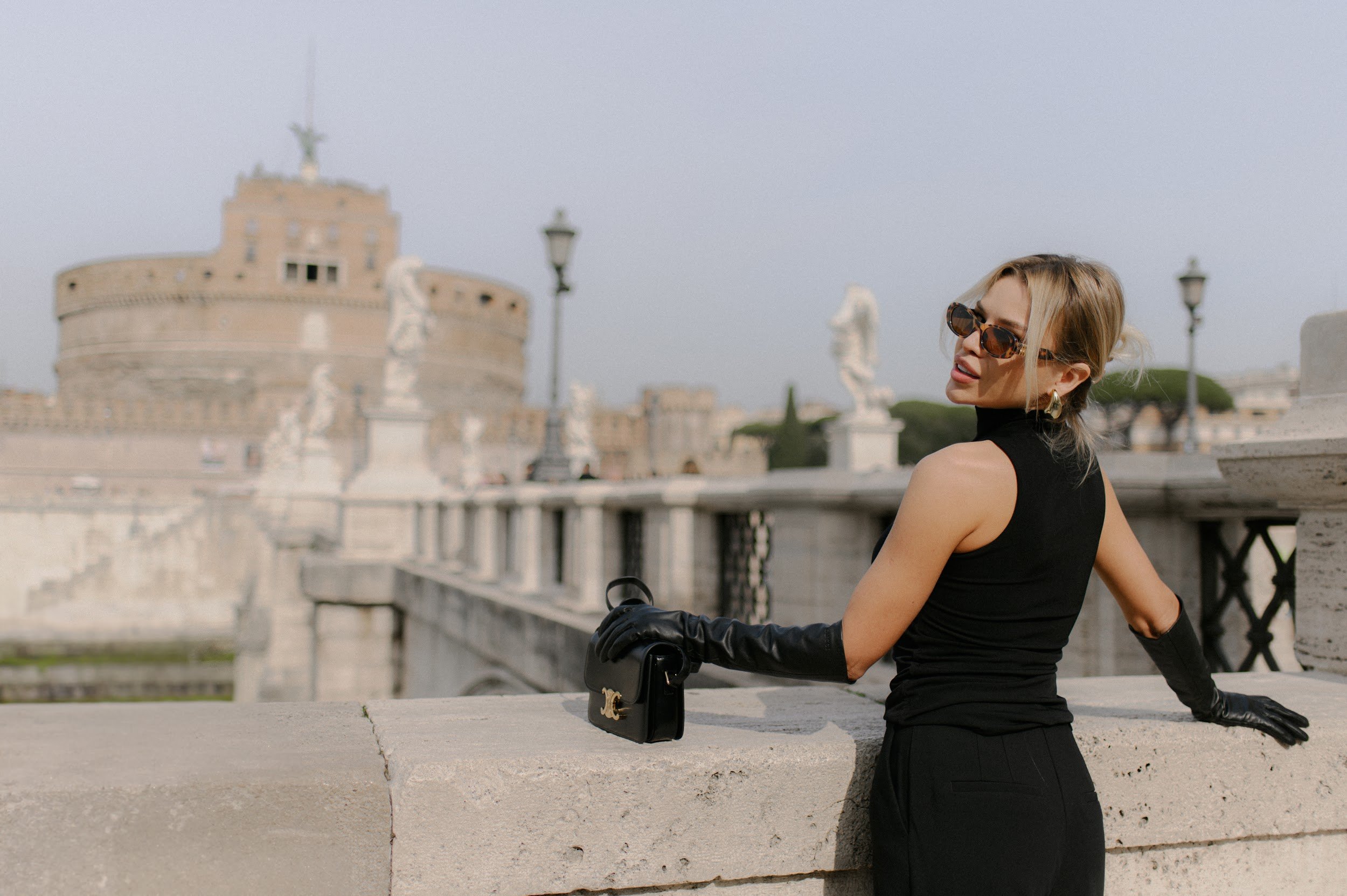 A woman wearing sunglasses and black gloves near Ponte Sant Angelo in Rome with Castel Sant Angelo in the background