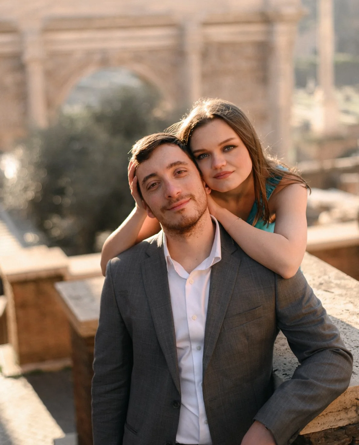 A young man in a gray suit and white shirt is sitting outdoors, with a young woman in a blue dress leaning on his shoulder and resting her head on his, both smiling softly against a blurred cityscape background with an arch bridge.