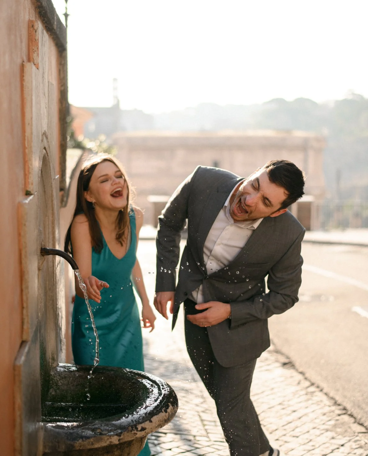 A woman in a teal dress and a man in a suit laughing and splashing water from a water fountain outdoors.