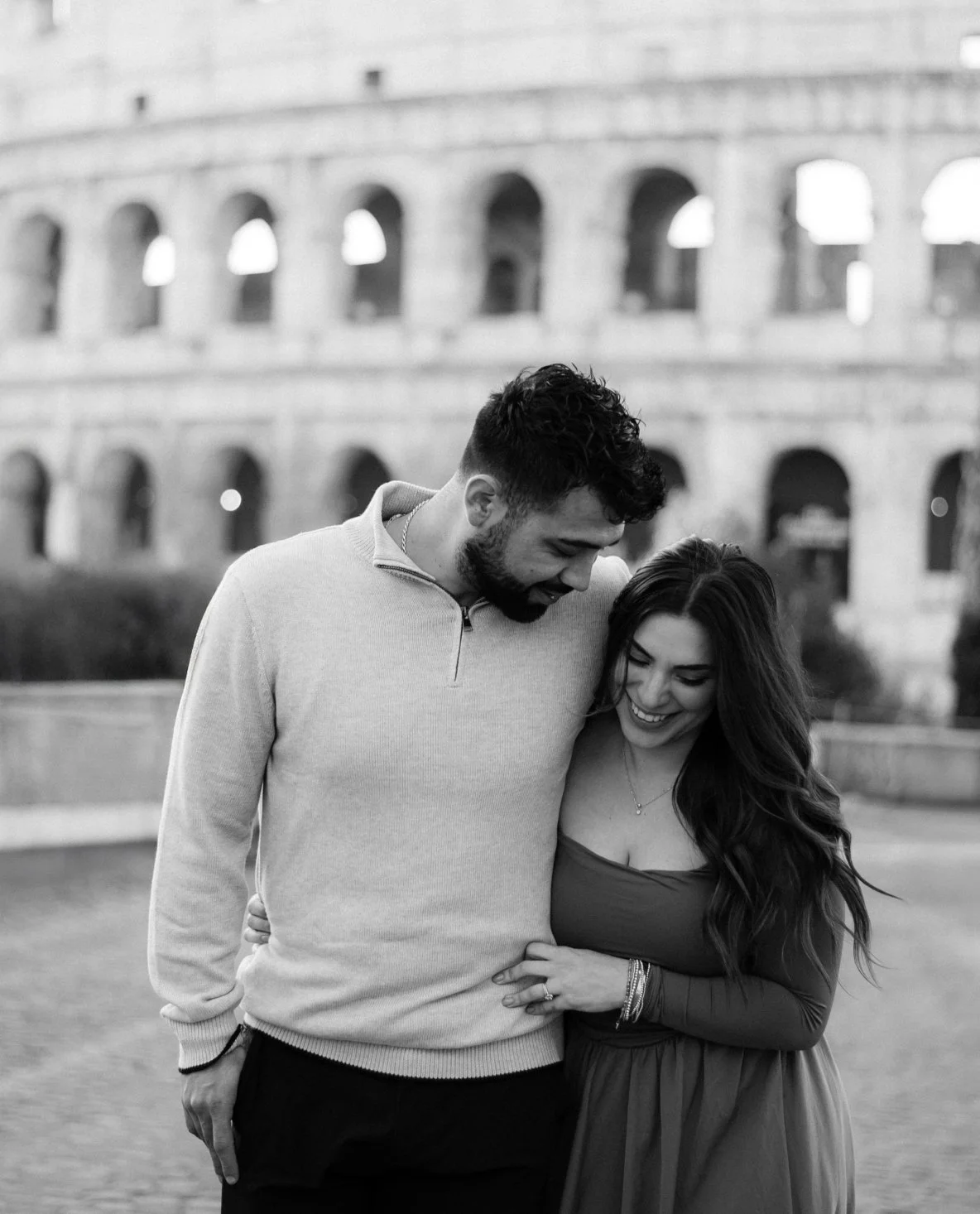 A man and woman smiling and hugging in front of the Roman Colosseum in black and white.