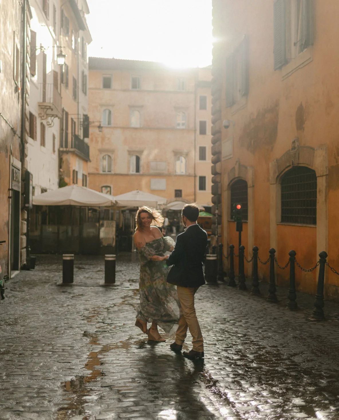 A couple dancing on a cobblestone street in a European city, with the woman wearing a floral dress and the man in a blazer, illuminated by sunlight.