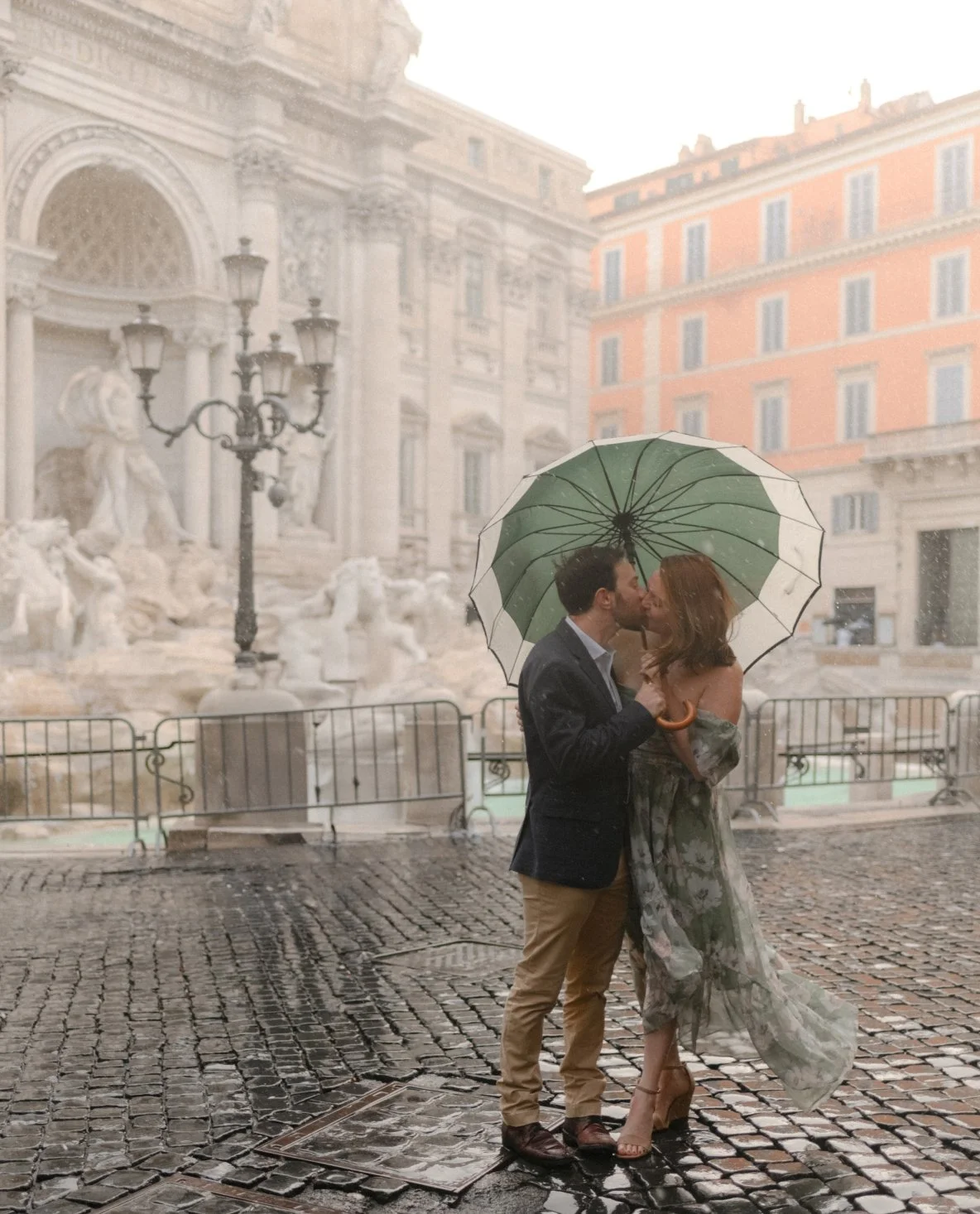 A couple kissing under a green and white umbrella on a rainy street, with historic buildings and sculptures in the background.