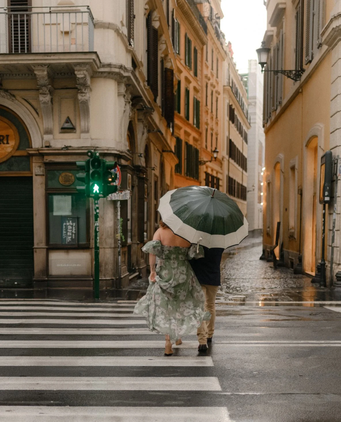 A couple walking across a city crosswalk in the rain, sharing a large umbrella. The bride is wearing a floral wedding dress, and the groom is dressed in beige pants and a dark jacket. The street is wet with reflections from the rain, and there are historic-style buildings and a traffic light showing a green pedestrian signal in the background.