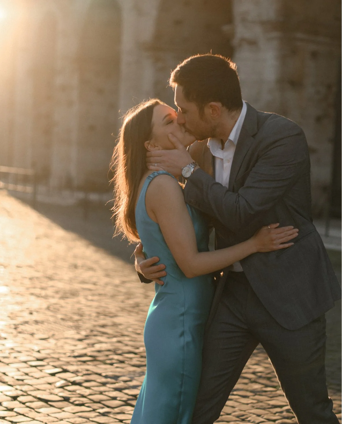A couple sharing a kiss outdoors during sunset, with a cobblestone street and brick wall in the background.