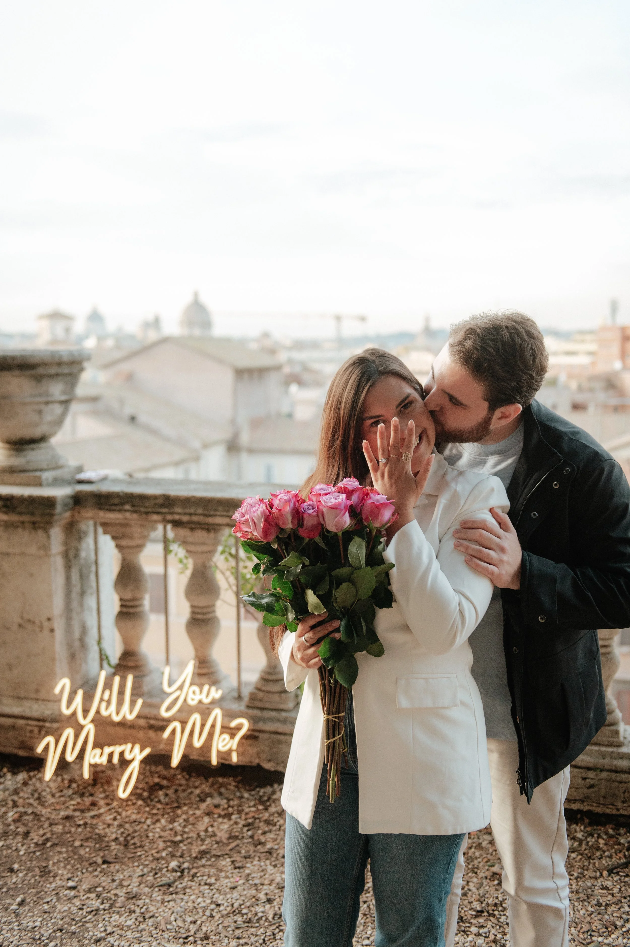 A man is kissing a woman on the cheek while she holds a bouquet of pink roses. The woman is smiling and showing her engagement ring, and the image features the text "Will You Marry Me?" in cursive writing.