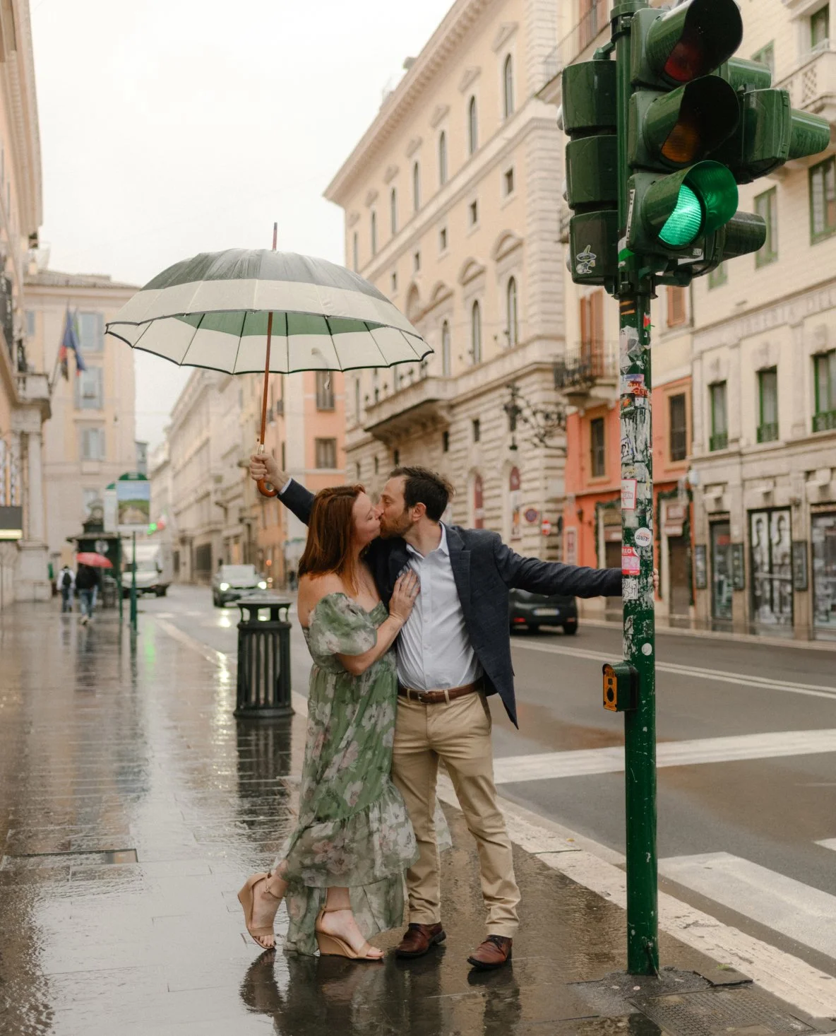 A couple sharing a kiss in the rain on a city street, with a woman holding an umbrella and a man leaning against a green traffic light pole.