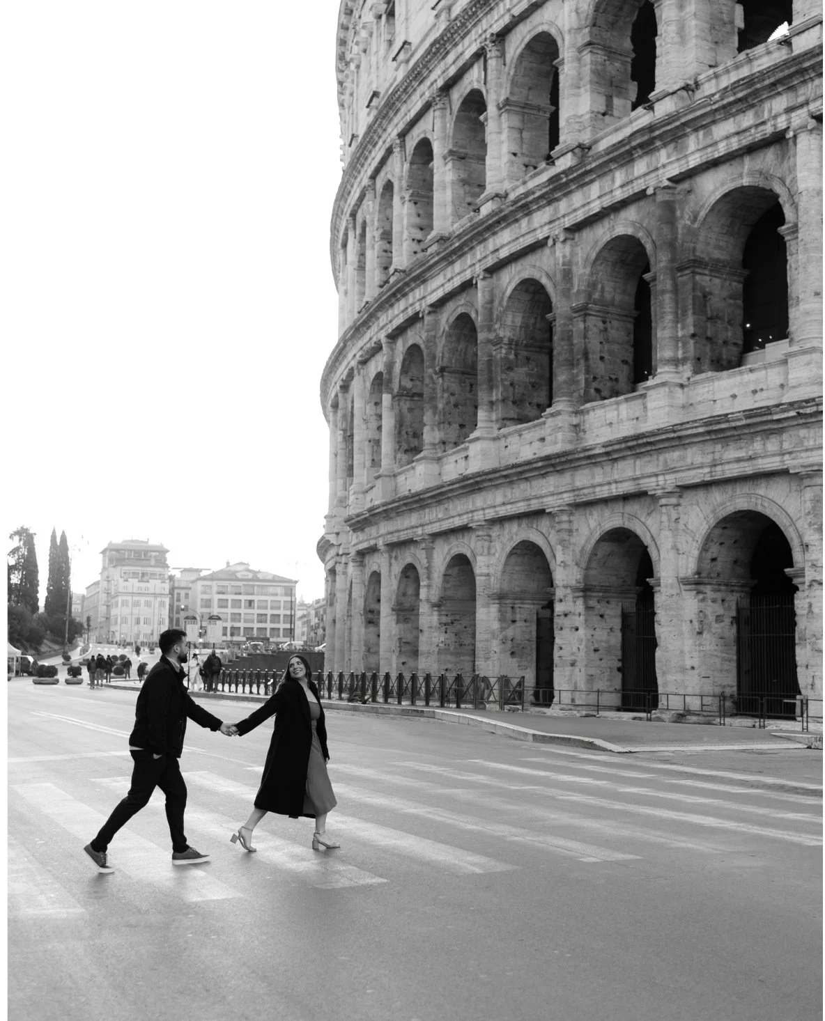 A couple holding hands and crossing a street near the Colosseum in Rome, Italy. The photo is in black and white.