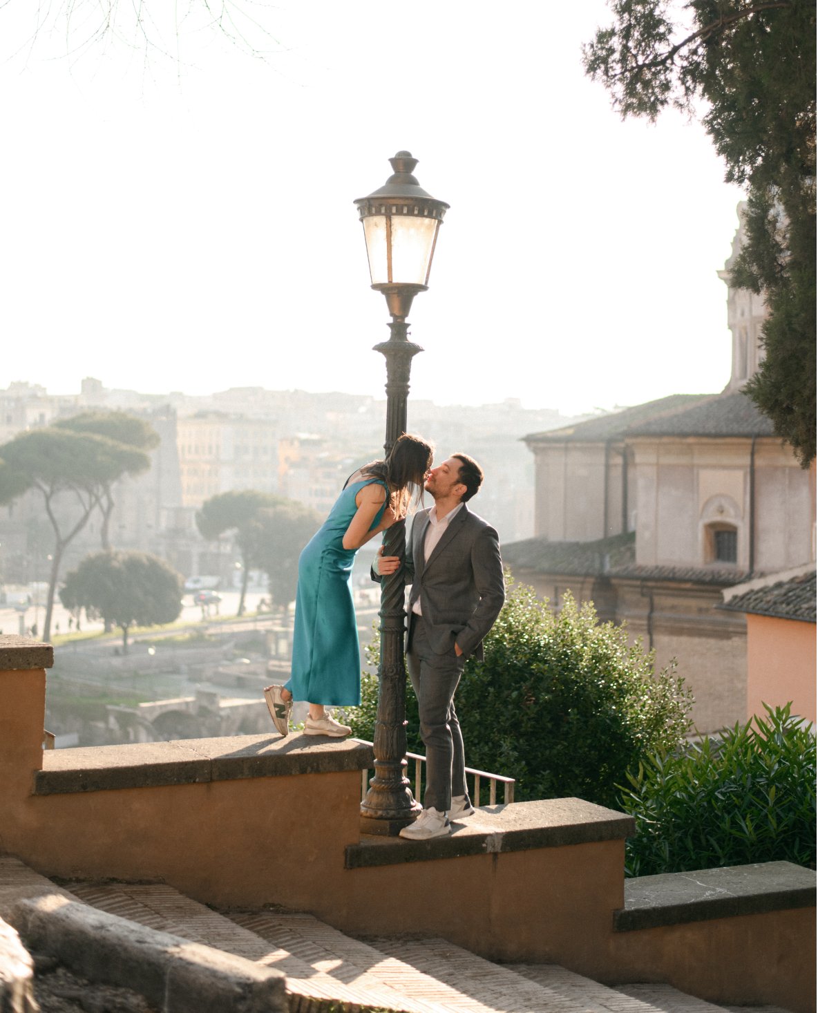 A woman in a blue dress is leaning over a street lamp to kiss a man in a gray suit. They are outdoors with a cityscape in the background.