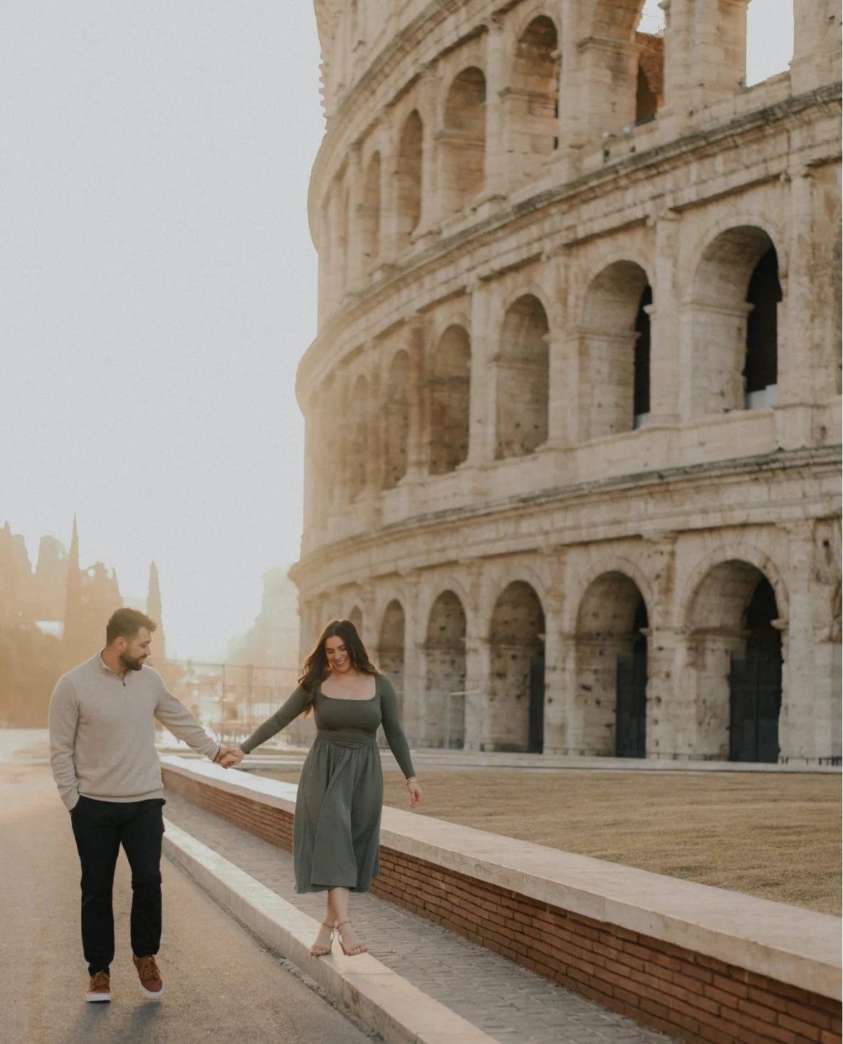 A couple walking hand-in-hand near the Colosseum in Rome at sunset, with the woman balancing on a low wall.