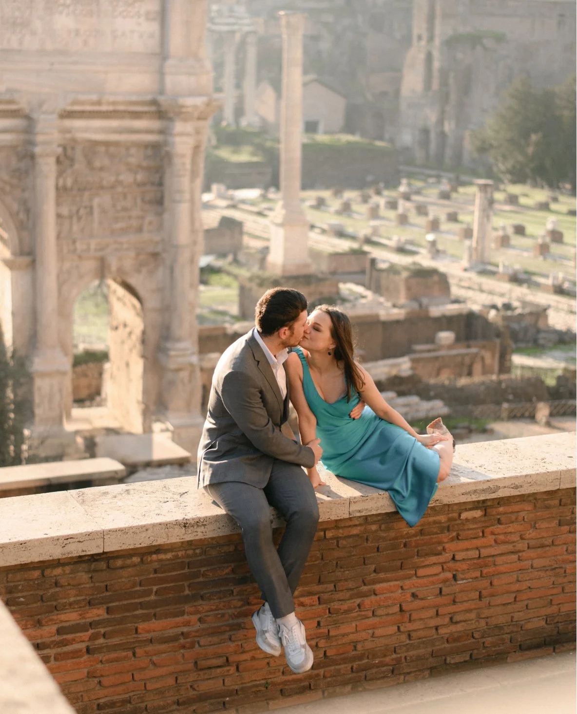 A man and woman sitting on a brick ledge, kissing, with an ancient ruin and cityscape in the background.