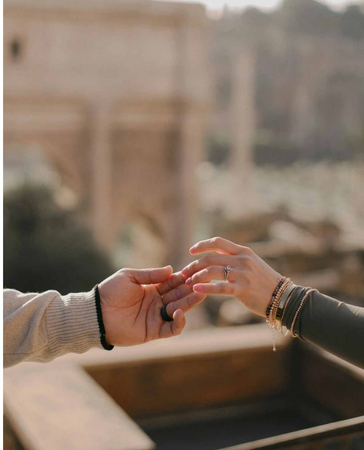 Two hands reaching out and touching each other, one with multiple bracelets and a ring, the other with a ring, against a blurred outdoor background.