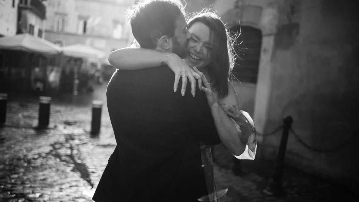 A black-and-white photo of a woman and a man hugging and smiling on a rainy street, with blurred buildings and umbrellas in the background.