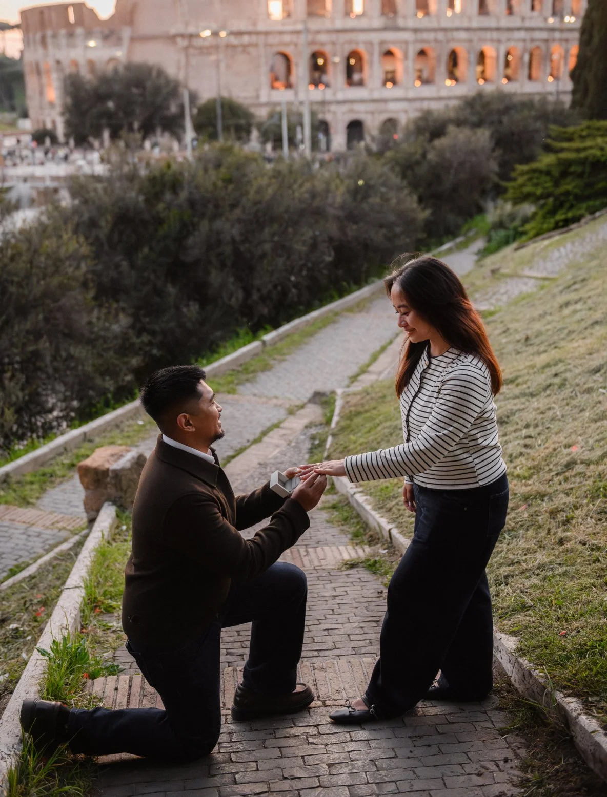 A man kneeling on one knee holding a small box, presenting a ring to a woman who is smiling and reaching out to take it, on a brick pathway with greenery and the Colosseum in the background.