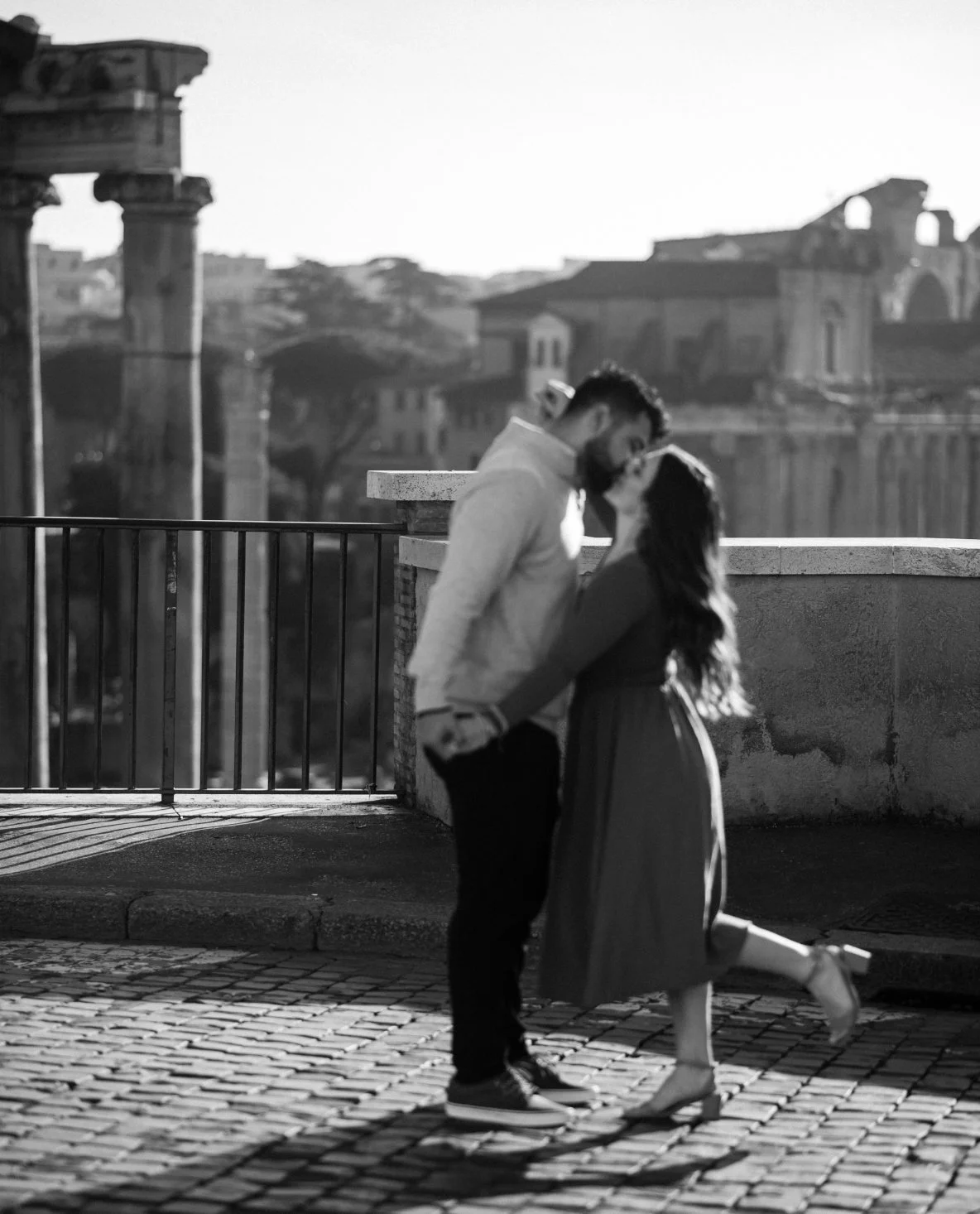 A black-and-white photo of a couple standing close on a cobblestone street, leaning in for a kiss, with historic ruins in the background.