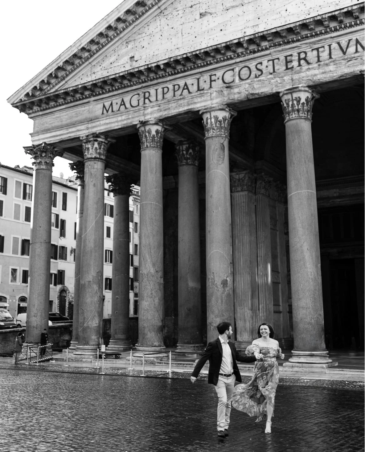 A black-and-white photo of a historic building with large columns and a pediment inscribed with Latin text. Two people are walking and holding hands in front of the building, with the woman wearing a floral dress and the man in a blazer and pants.