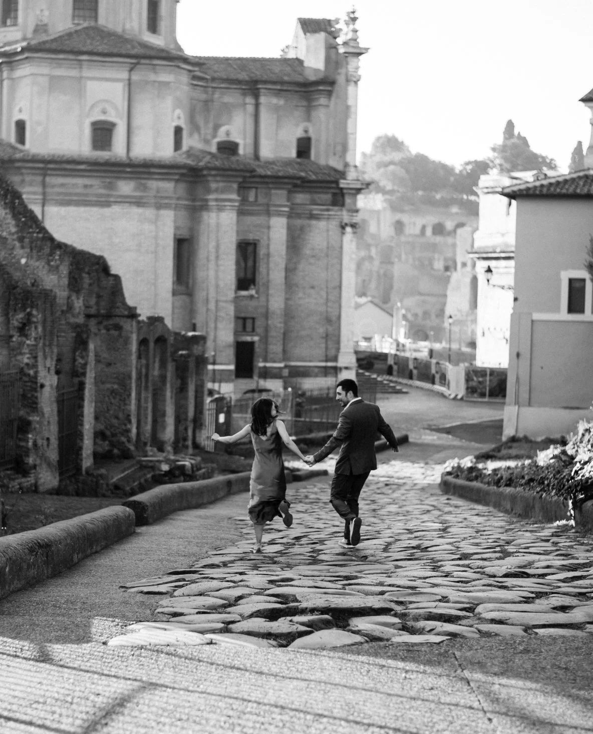 A black and white photo of a couple running hand in hand on a cobblestone street in an old European city, with historic buildings and church in the background.