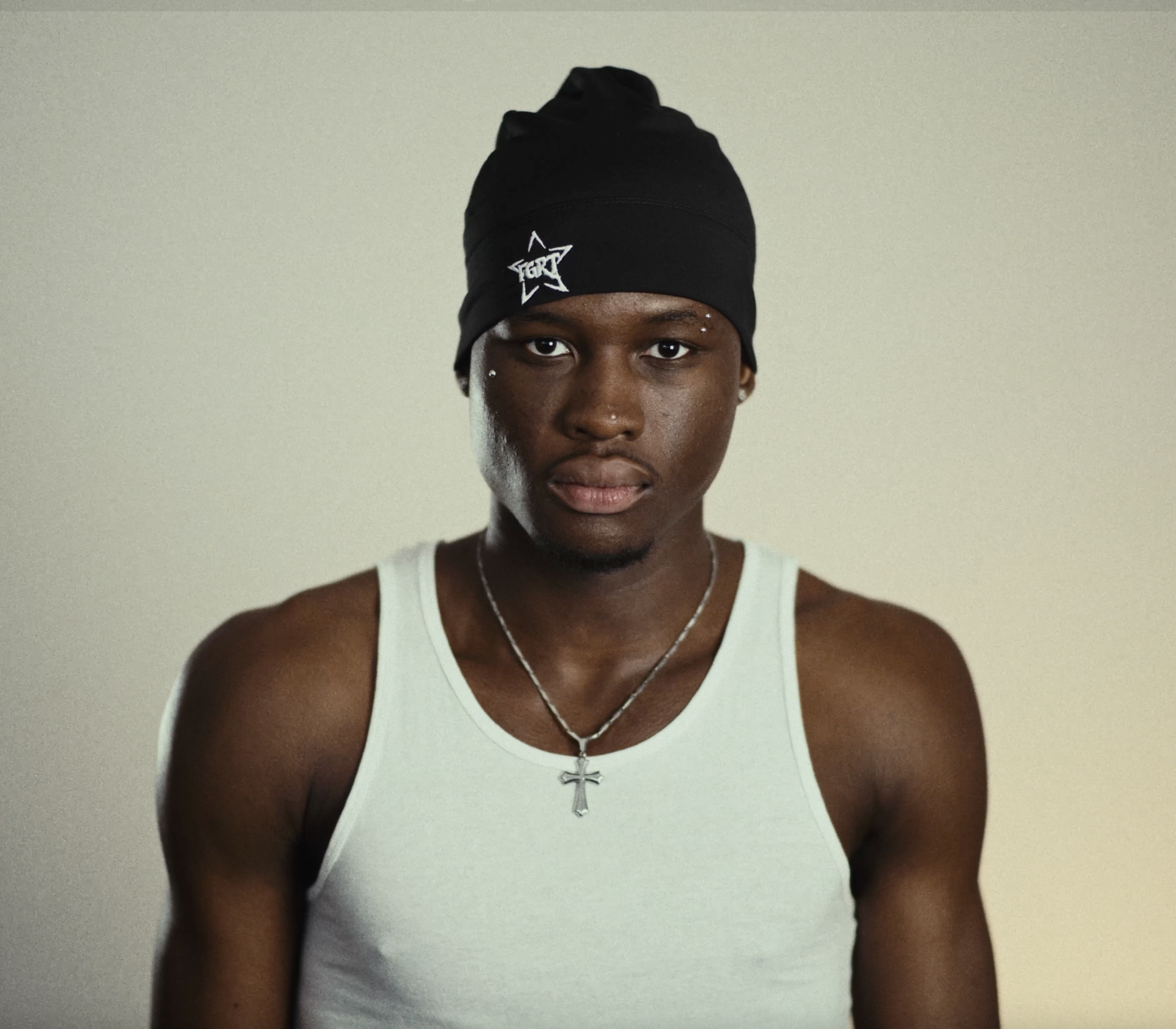 A young man wearing a black beanie hat, a white tank top, and a silver necklace with a cross pendant, standing against a plain background.
