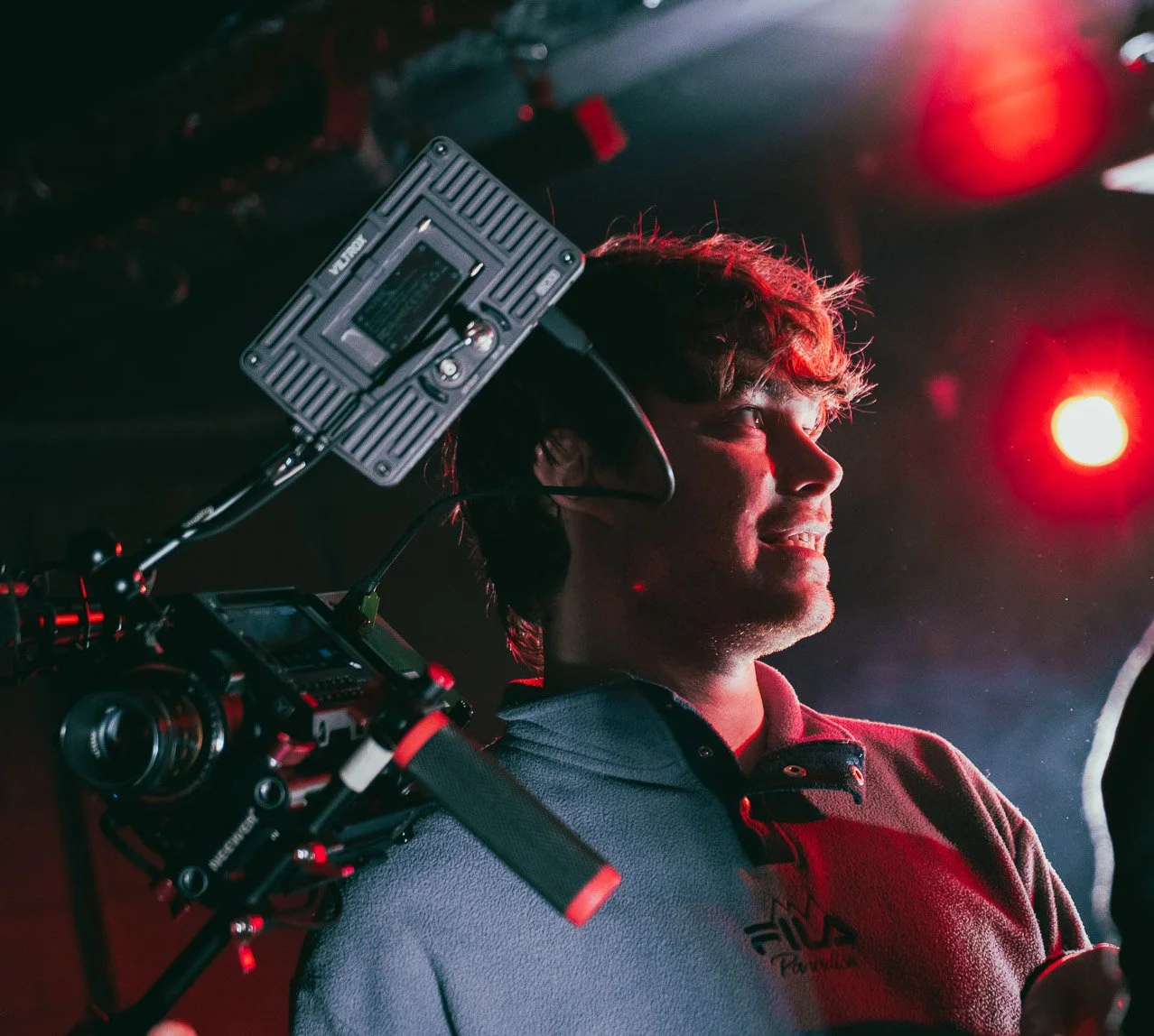 A young man with headphones smiling in a dimly lit space illuminated by red lights, with a professional camera mounted on a stabilizer nearby.