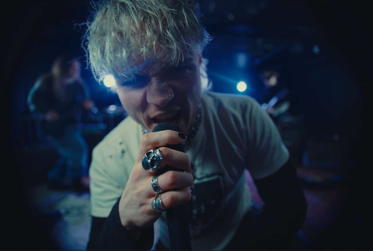 Person with short blonde hair singing into a microphone during a music performance, with band members playing in the background