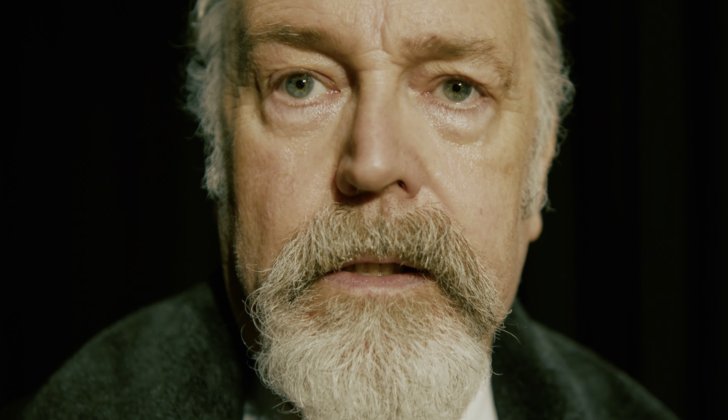 Close-up of an older man with a white beard and mustache, gray hair, blue eyes, and a serious expression, against a black background.