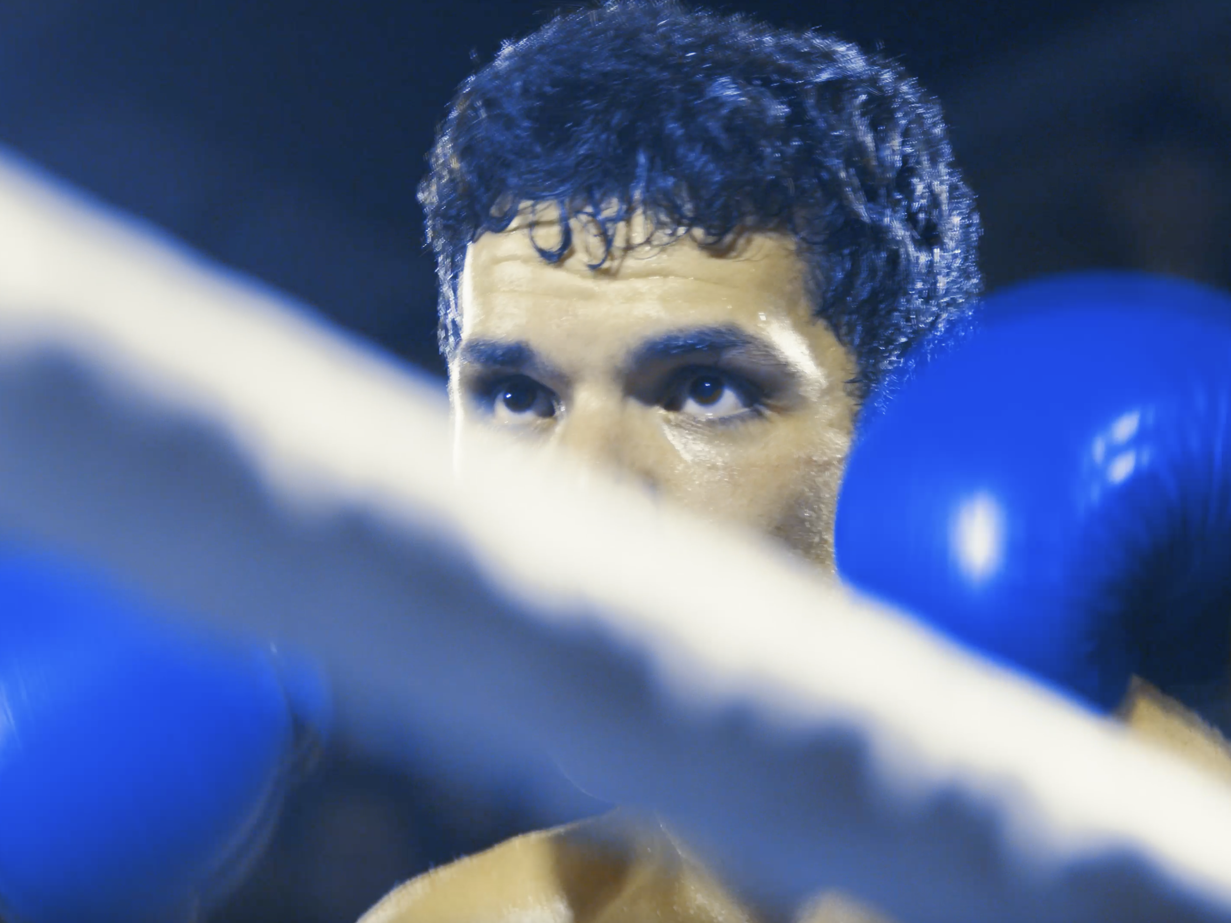 A male boxer with blue gloves looking with intense focus, possibly in a boxing ring.