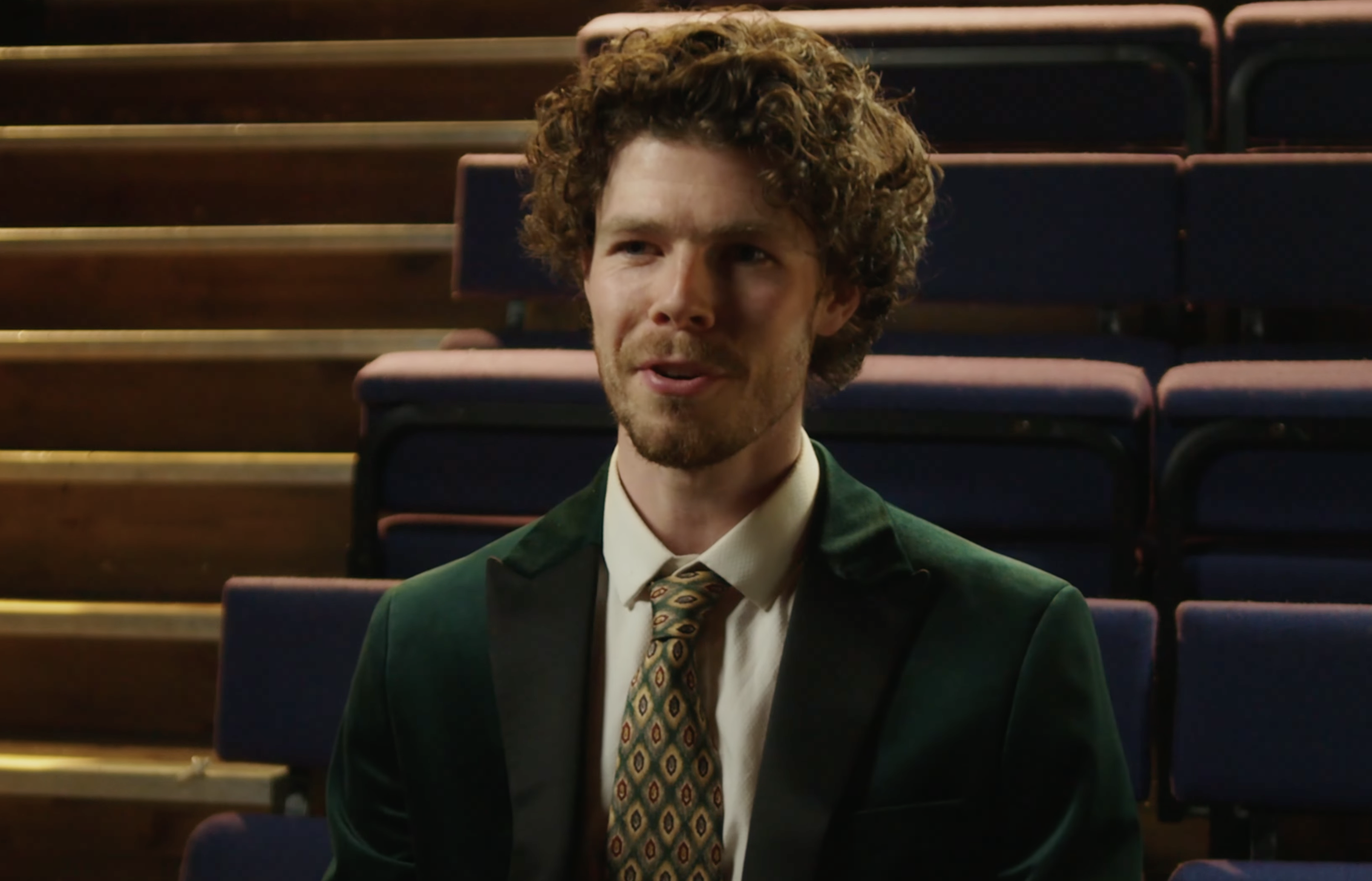 A man with curly brown hair and a beard wearing a green velvet blazer, white shirt, and patterned tie sitting in an auditorium with blue seats.