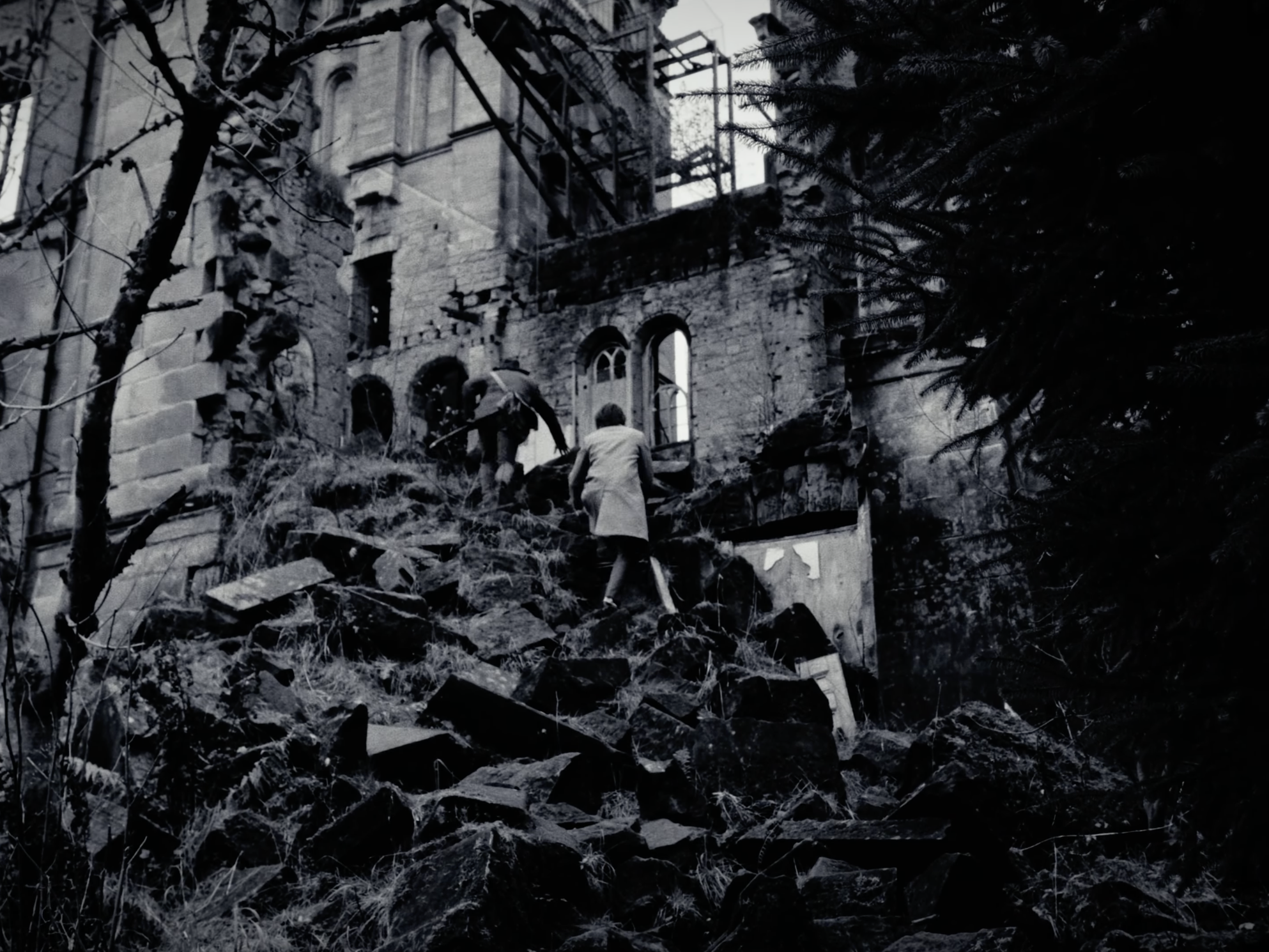 People walking on a rocky, debris-covered area outside a damaged and partially ruined building with arched windows.