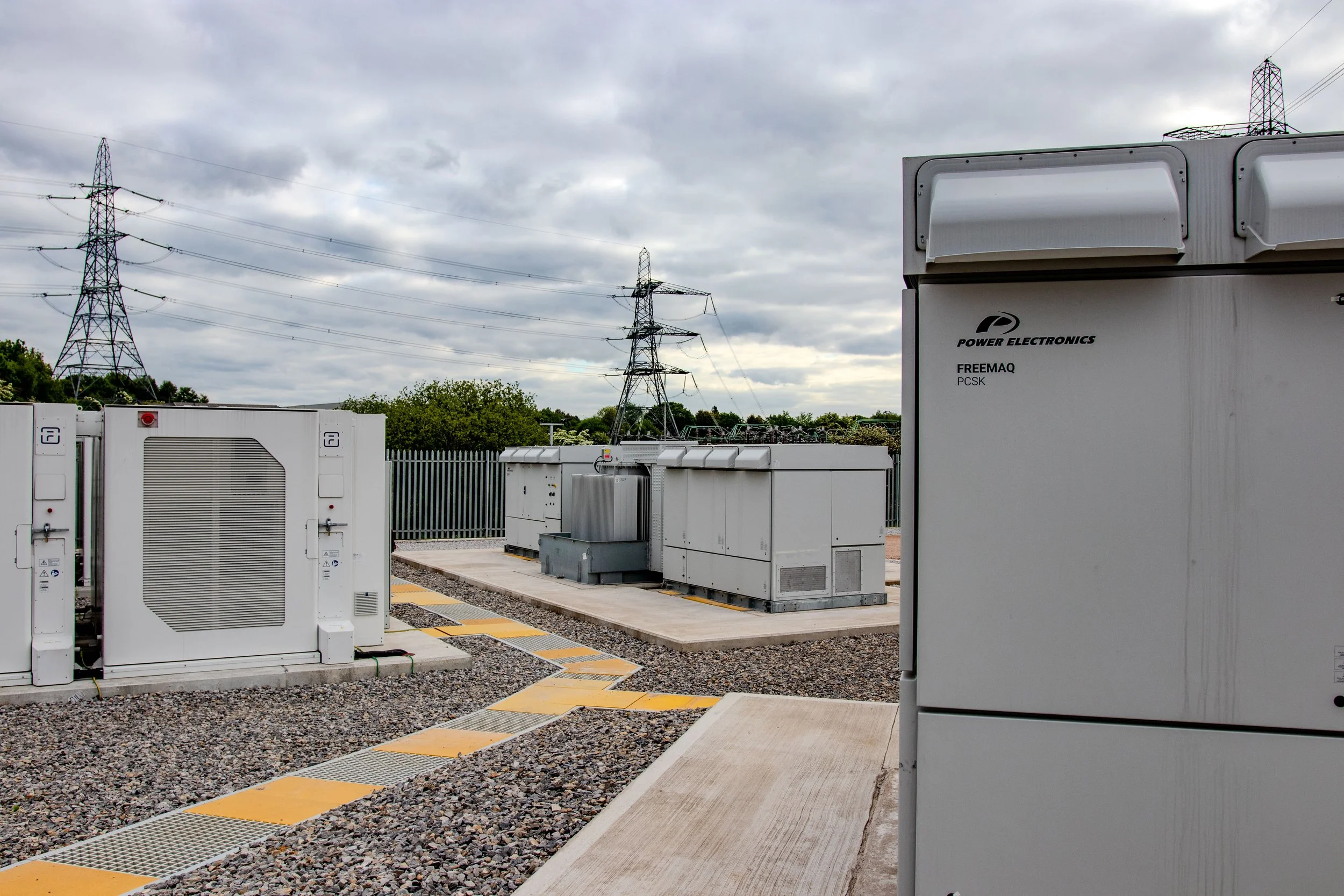 Electrical equipment and transformers outdoors at a utility site, with a paved walkway, gravel ground, and power lines and pylons in the background under a cloudy sky.