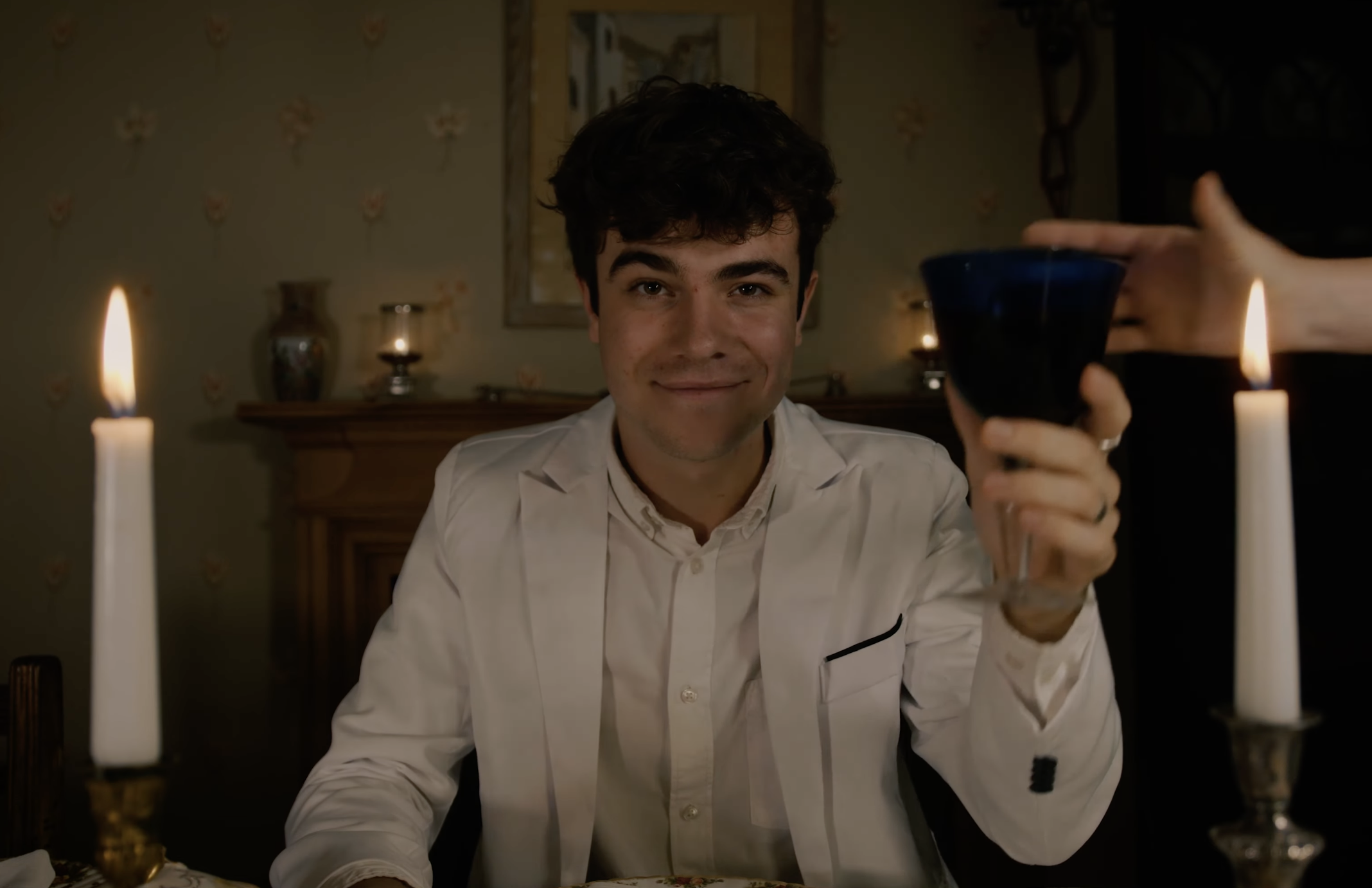 Young man in a white suit raising a glass at a candlelit dinner.