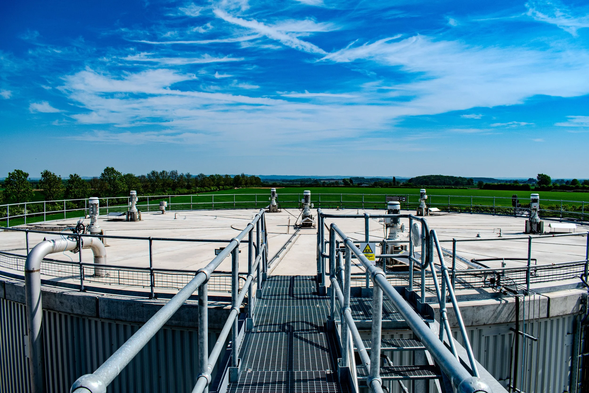 View of a large industrial water or chemical storage tank with metal walkways and railings, set against a green rural landscape and blue sky with scattered clouds.