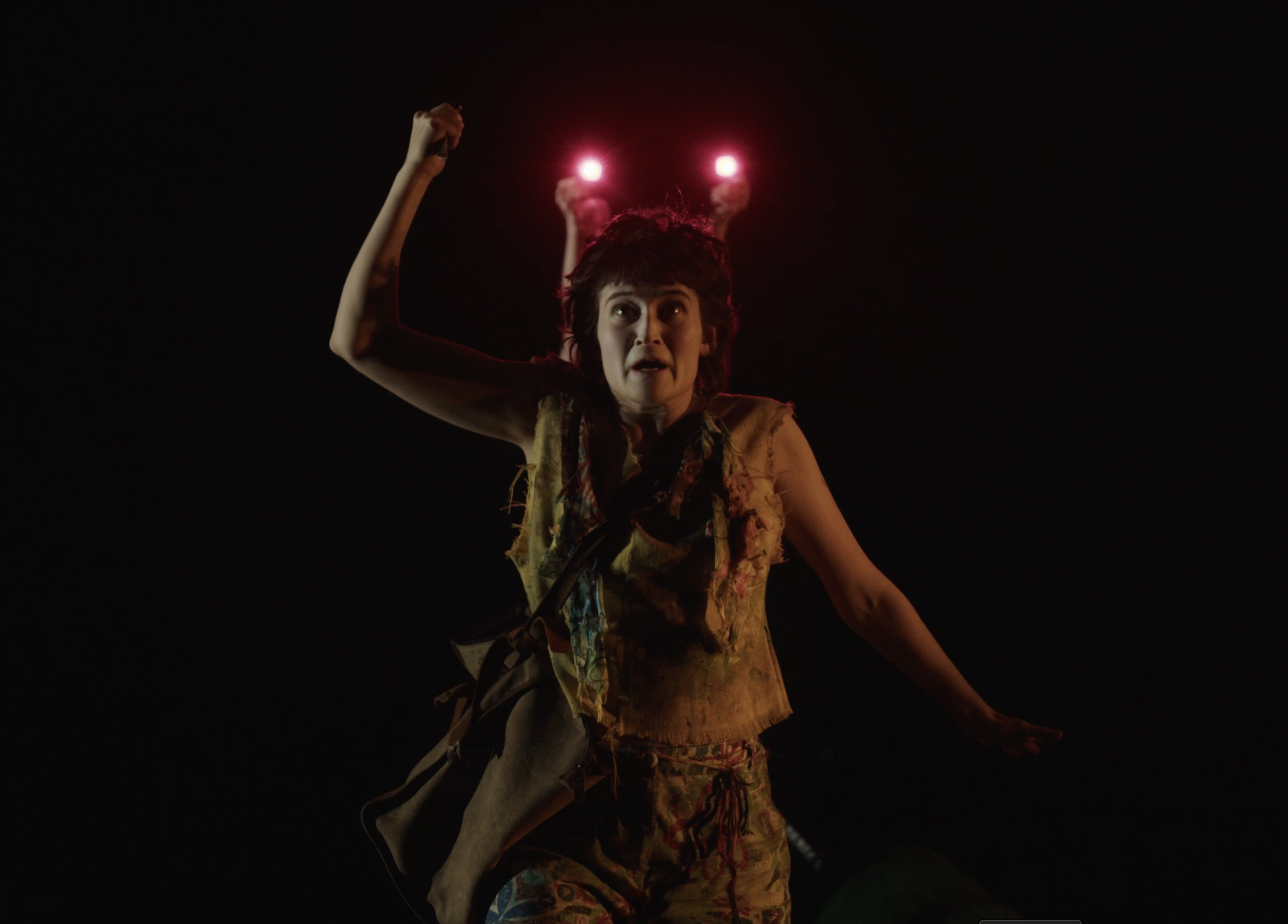 A woman on stage performs, dressed in tattered clothing with arms raised and an intense expression. Red stage lighting shines behind her.