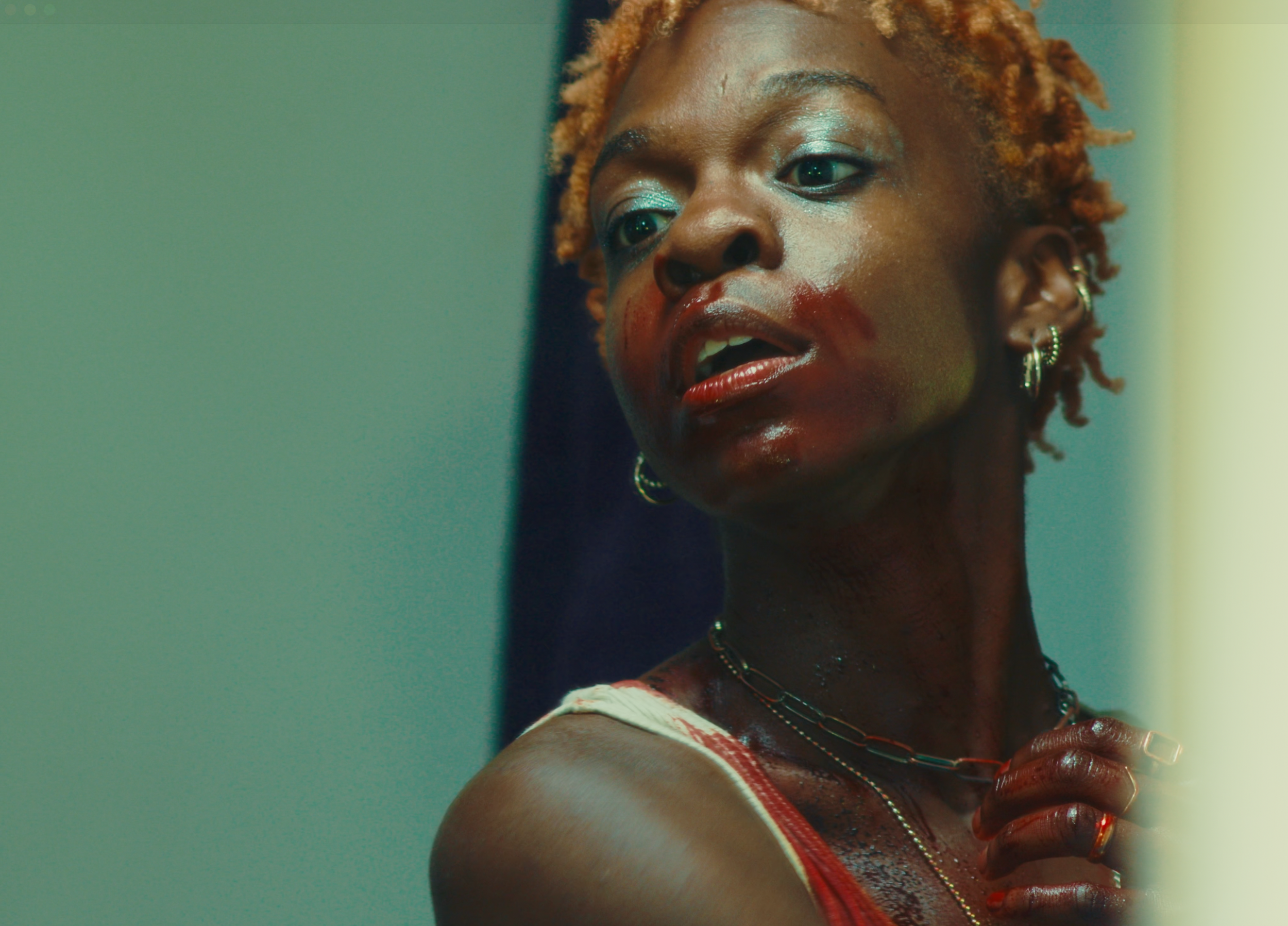 A woman with short, curly orange hair, wearing earrings, a chain necklace, and a red top, looking to her right with a serious expression.