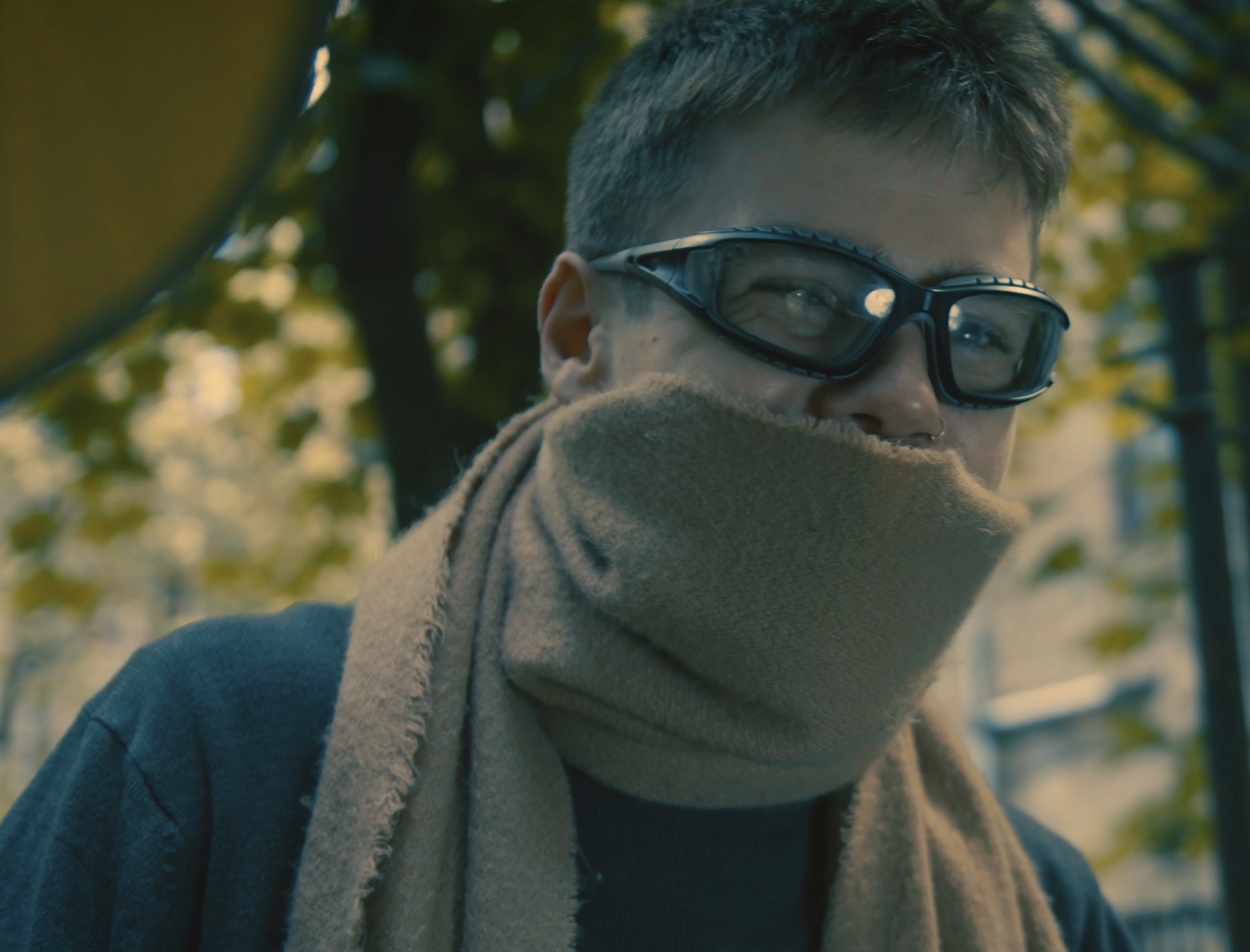 A person wearing goggles and a brown scarf covering the lower face, outdoors with blurred trees in the background.