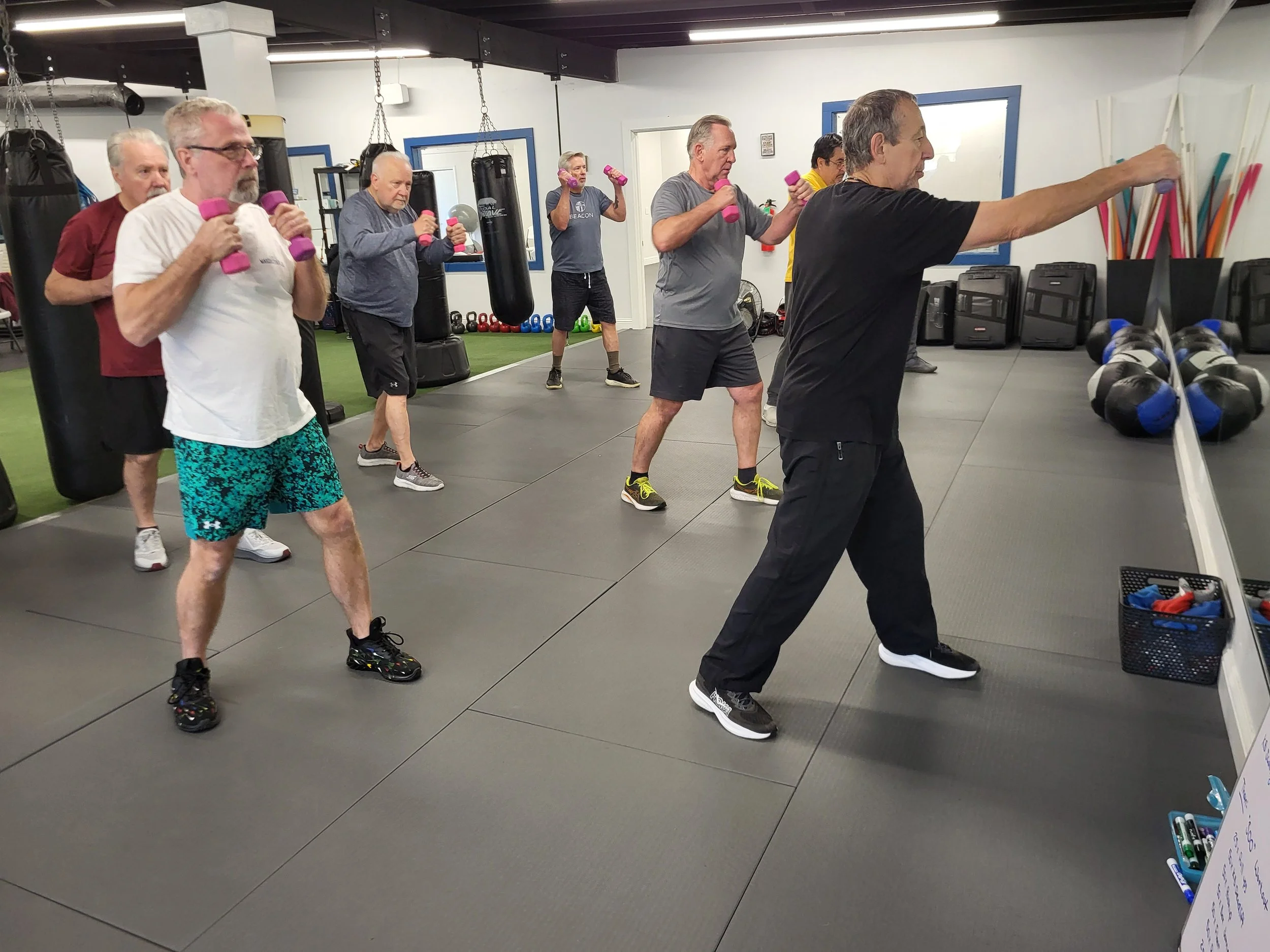 Group of older adults participating in a fitness class, lifting pink dumbbells and following a trainer in a gym with punching bags and exercise equipment.