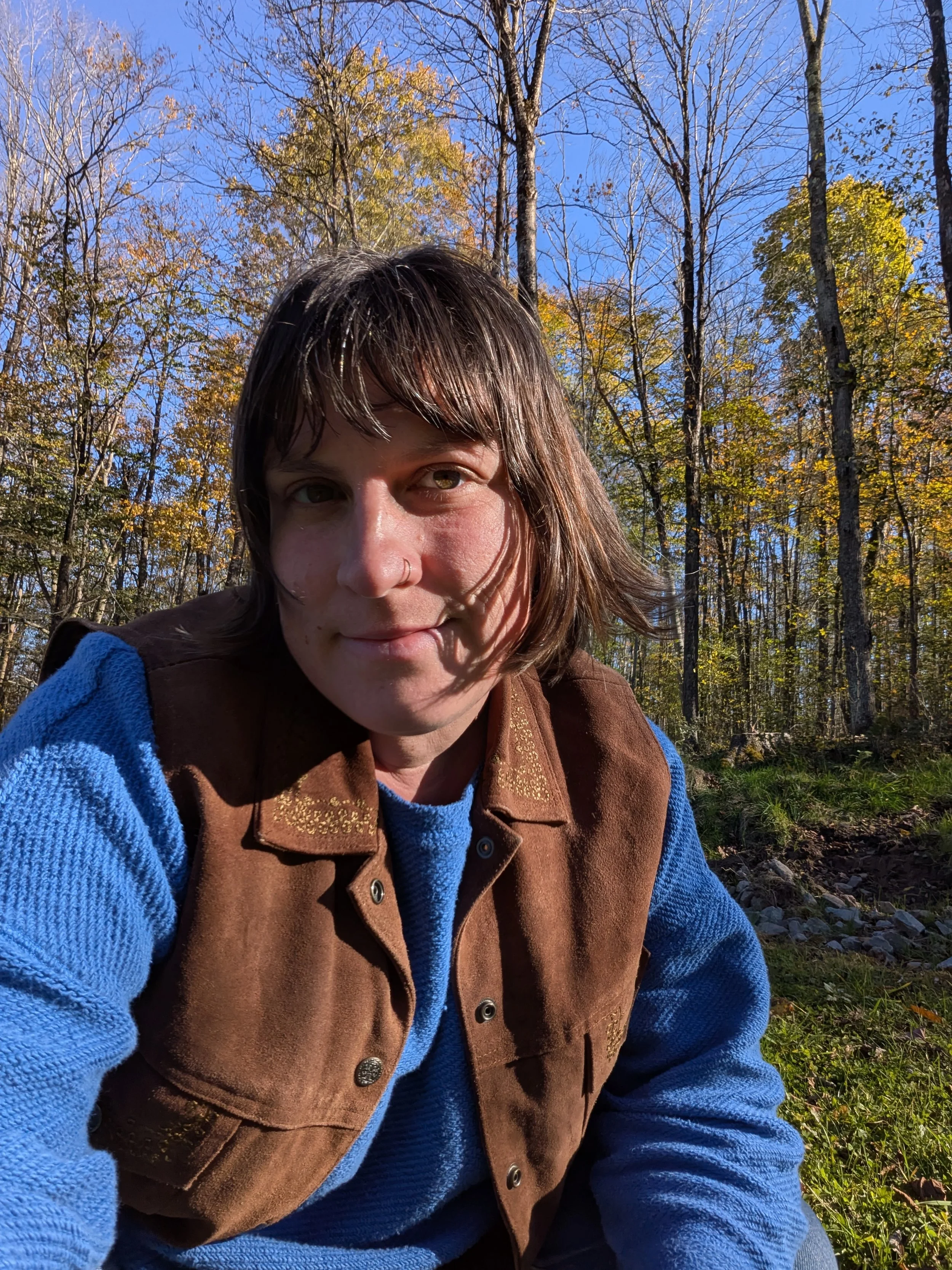 Image shows Nora, Mossy Hollow's founder wearing a blue shirt and brown vest smiling and looking into the camera while sitting in the forest with trees in the background that have changed colors in the fall