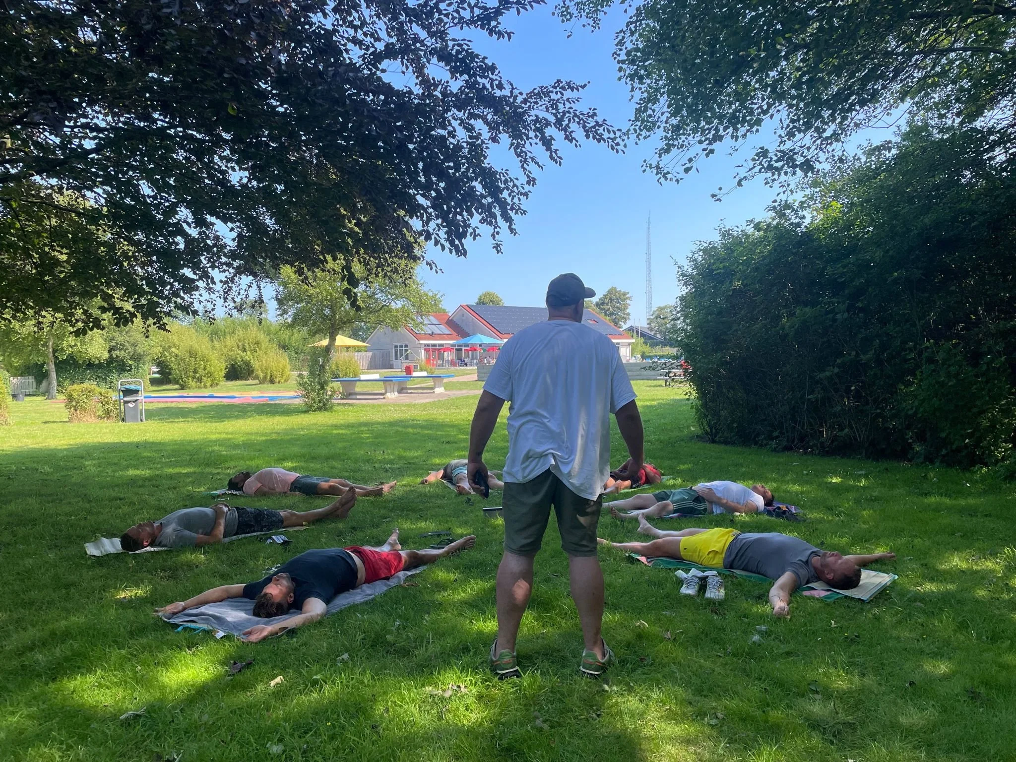 Mensen liggen op matten op het gras tijdens een yoga sessie in een park, onder schaduwrijke bomen, met een instructeur die toezicht houdt.
