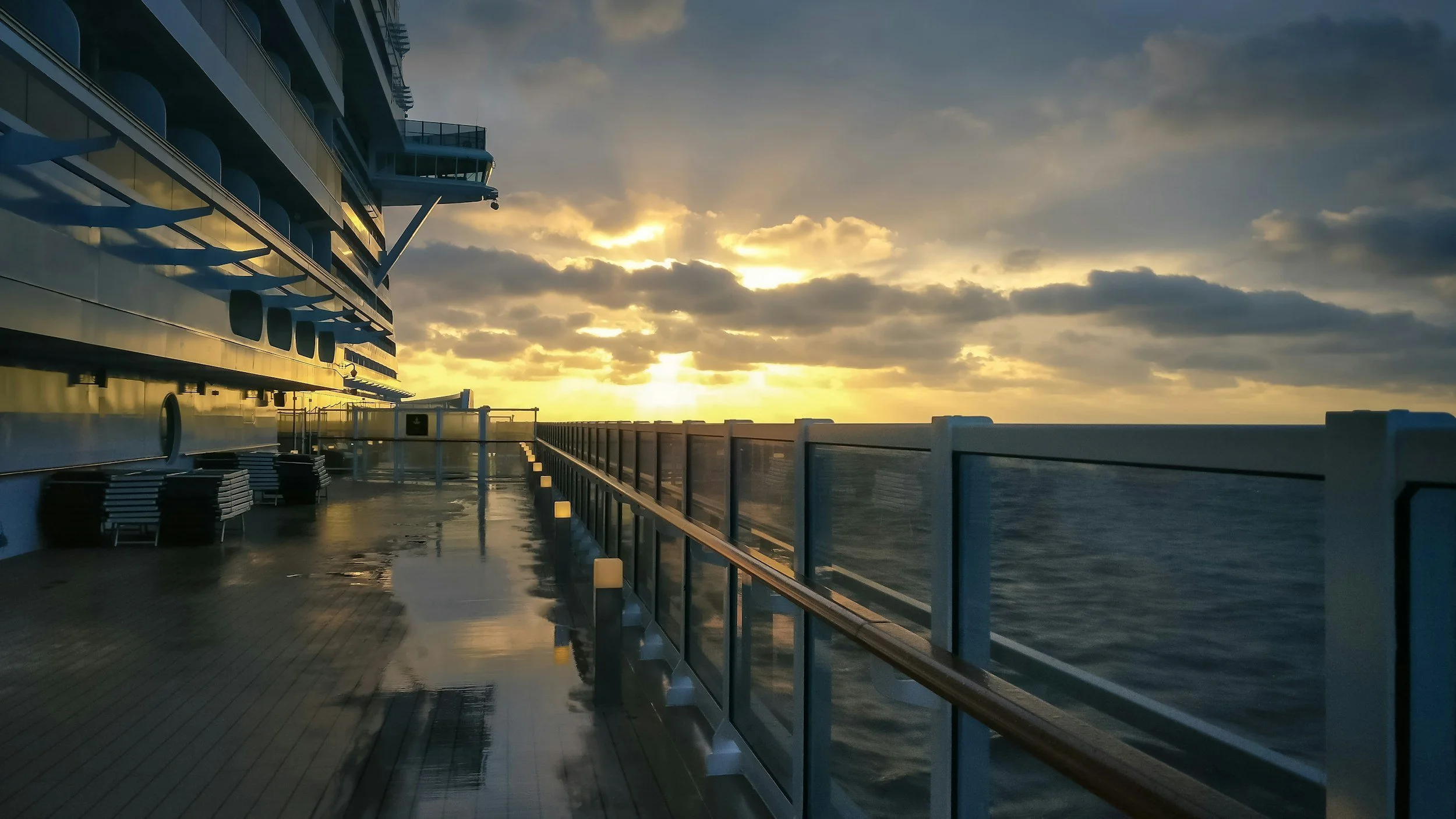 View of the deck of a cruise ship at sunset with chairs and a railing overlooking the ocean.
