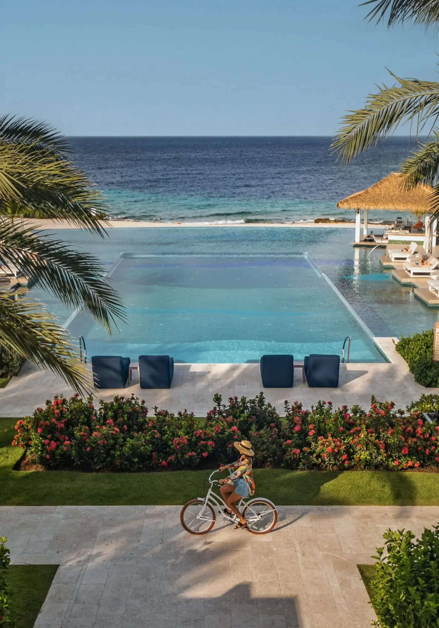 A young girl riding a bicycle on a paved pathway near a colorful garden of flowers. In the background, there's a large swimming pool, loungers, and a thatched-roof cabana overlooking the ocean. Palm trees frame the scene, and the sky is clear.