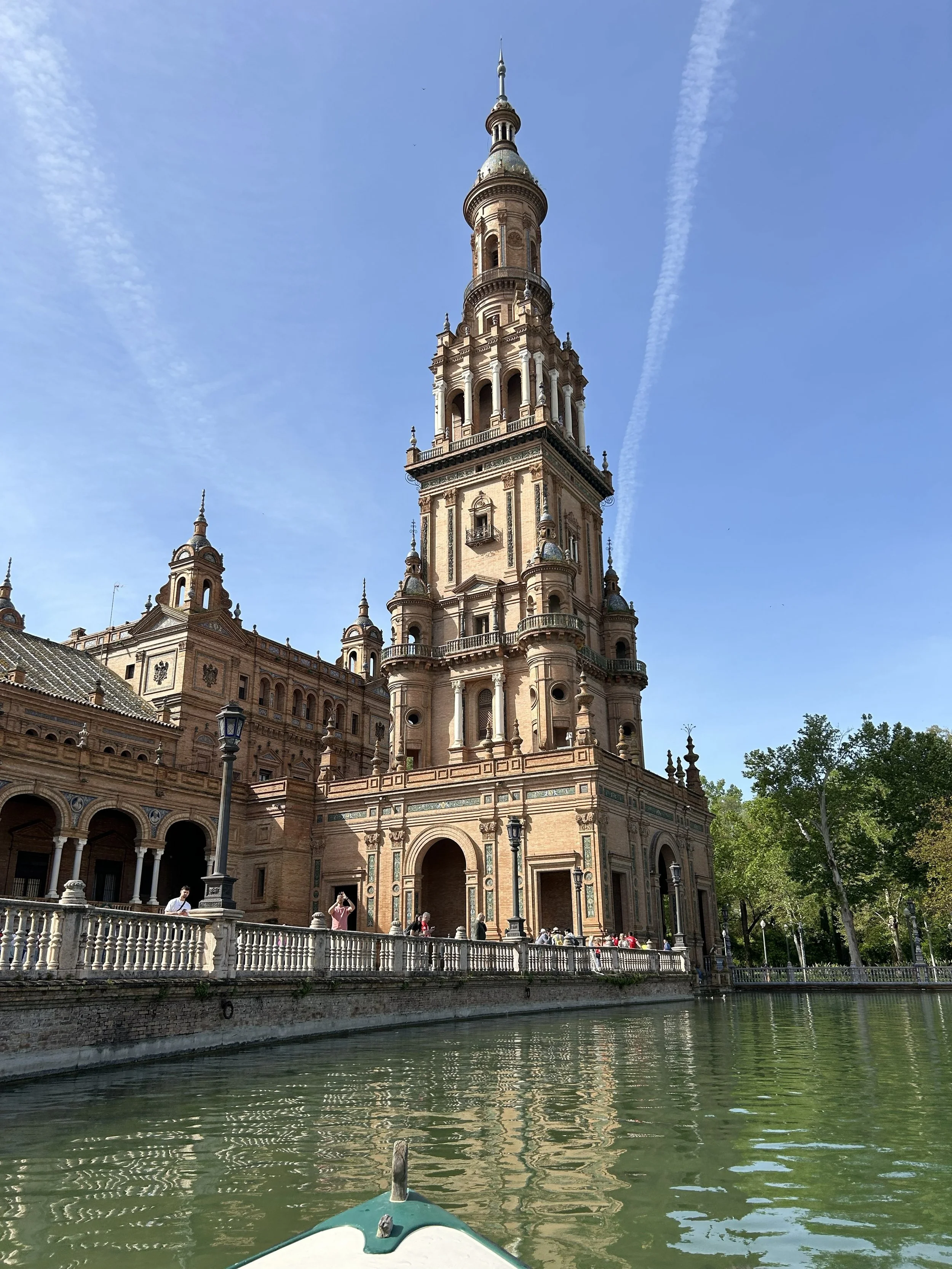 View of a historic castle-like building with a tall tower, seen from a boat on a river with green water. The building features ornate architecture with spires, decorative windows, and details in a warm stone color. People are visible on the promenade in front of the building, and trees are on the right side of the image.