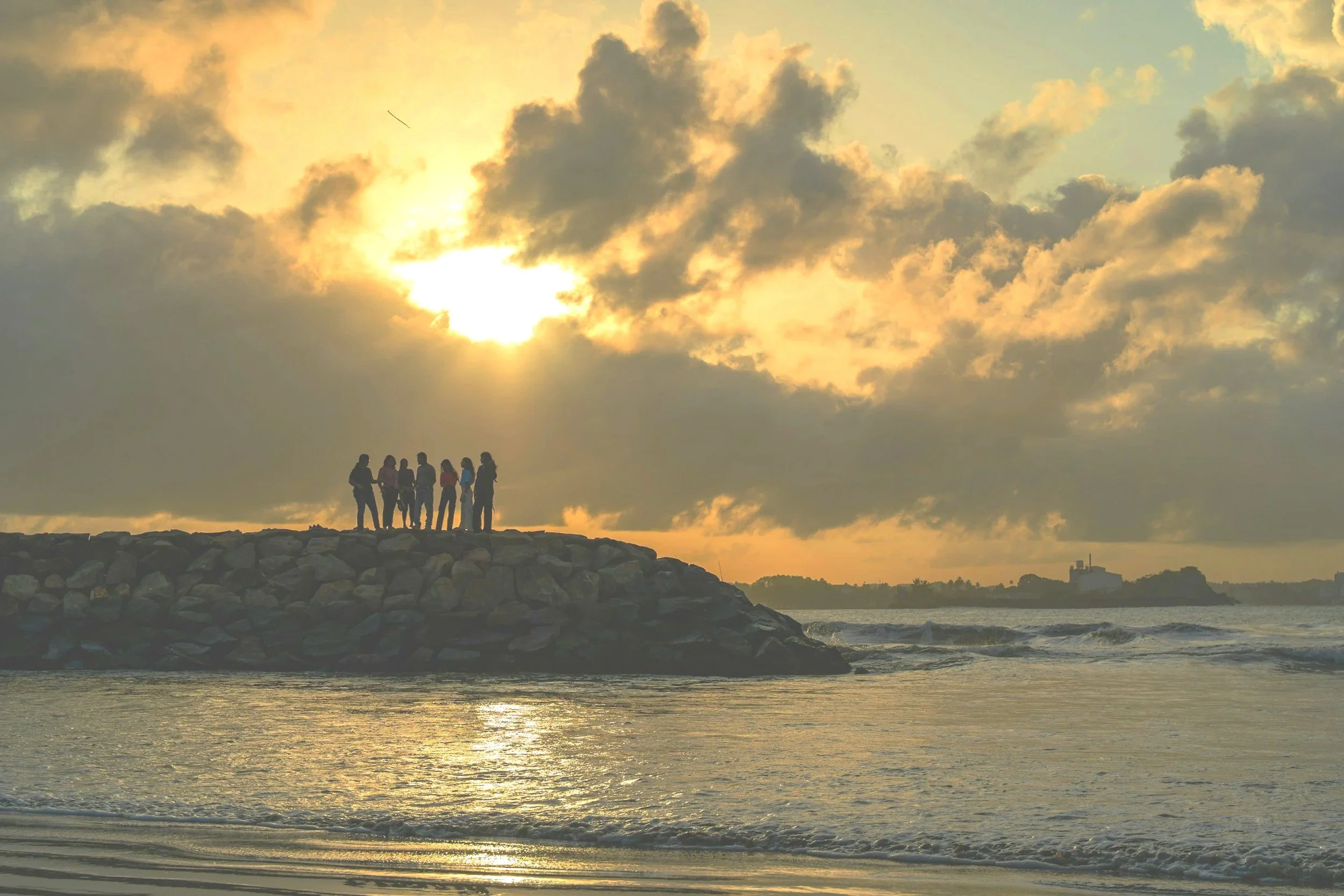 A group of people standing on rocks at the edge of the water during sunset, with a cloudy sky and a small building in the distance.