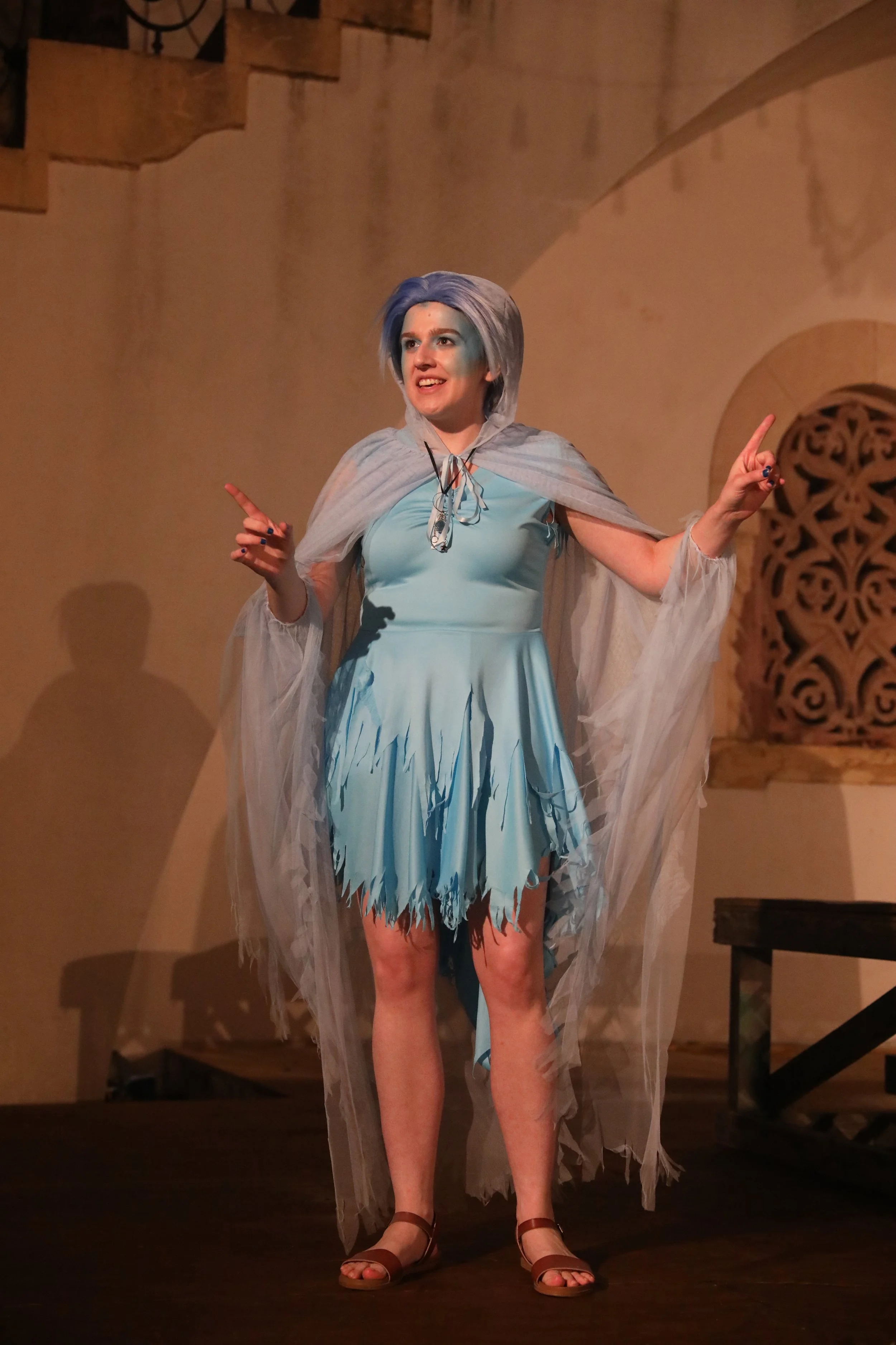 Woman (Katherine Eichenberger) dressed in a light blue, torn dress with a sheer cape, with blue hair, standing on a wooden stage, smiling and gesturing with her hands.